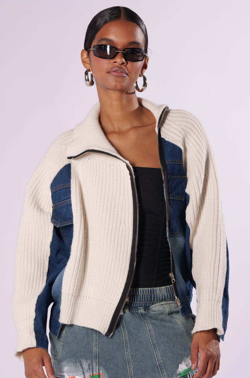 A woman wearing the "SOMETHING ABOUT YOU SWEATER" cream and denim jacket over a black top stands against a plain background. She accessorizes with dark sunglasses, hoop earrings, and dark wash denim jeans with patches. The jacket is partially unzipped, revealing the top underneath.