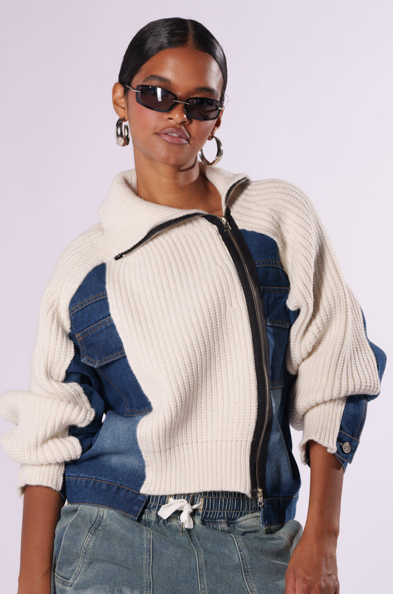 A woman wearing sunglasses and hoop earrings stands confidently in her stylish outfit. She sports a braided hairstyle and is dressed in the SOMETHING ABOUT YOU SWEATER, featuring a cream and blue knit zip-up jacket with denim accents, paired with loose-fitting blue-gray pants. The background is plain light grey.