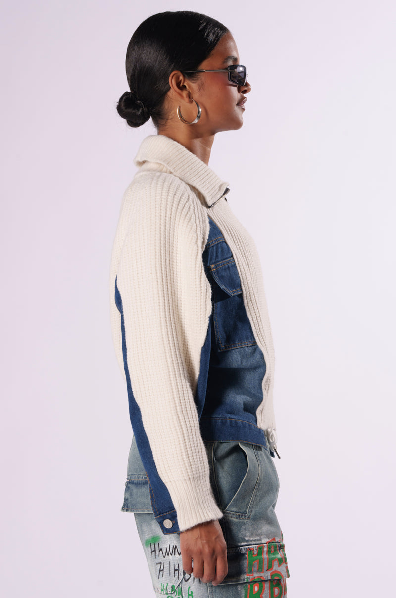 A person with dark hair styled in a low bun is shown in profile. They wear dark sunglasses, a SOMETHING ABOUT YOU SWEATER featuring beige fabric and blue denim patches, and jeans with colorful embroidered text on the dark wash denim. The background is plain and light-colored.