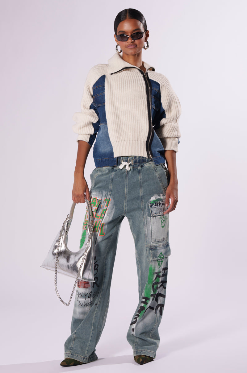 A person stands confidently against a plain background, sporting the SOMETHING ABOUT YOU SWEATER with its woven knit base. They pair it with wide-leg jeans adorned in colorful graffiti-style designs and complete the outfit with pointed-toe shoes. Their look is accessorized with sunglasses and a silver handbag.
