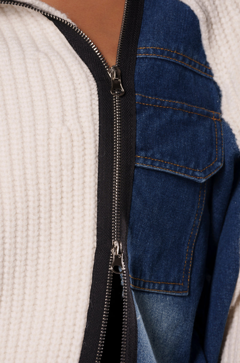 Close-up of a garment featuring a zipper. The left side of the zipper shows a white, ribbed fabric from the SOMETHING ABOUT YOU SWEATER, while the right side displays dark wash denim with pocket and seam details, creating a contrast between textures and colors.