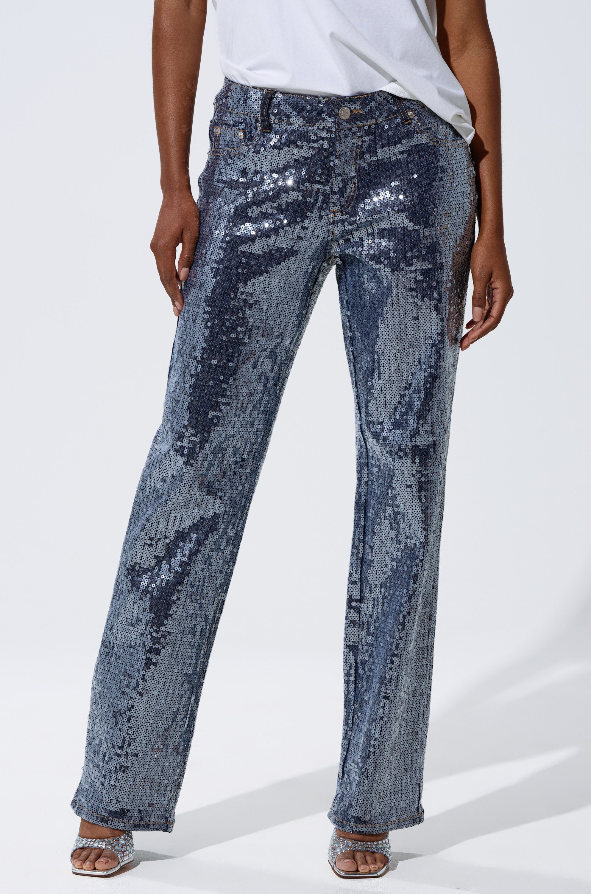 The lower half of a person in the ASHTON SEQUIN DENIM PANT, paired with a white shirt and open-toed heels, is shown against a plain white background.
