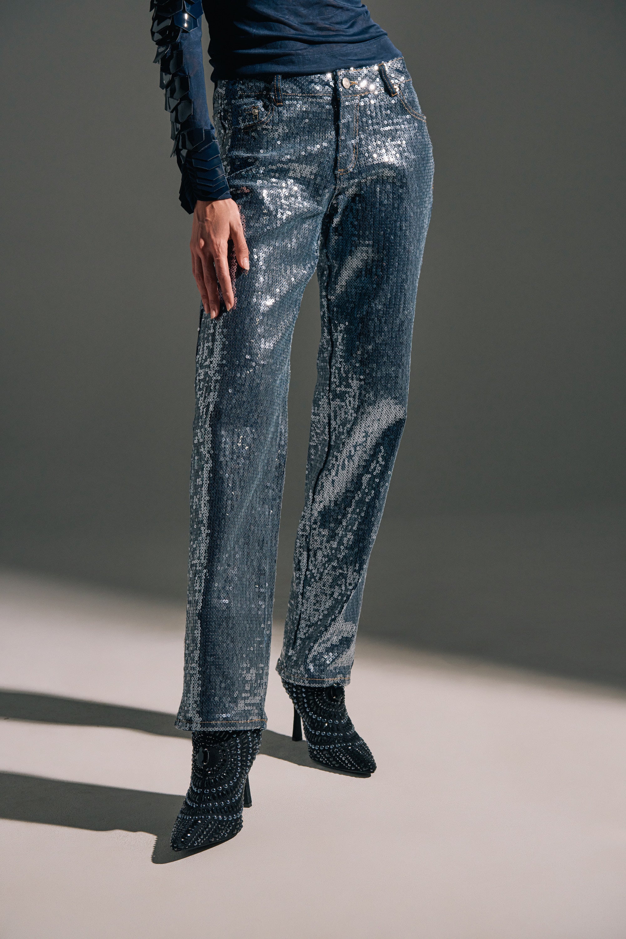 A person wears the ASHTON SEQUIN DENIM PANT paired with sparkly high-heeled boots and a long-sleeve navy top. The image, cropped from the chest down, features a neutral background.