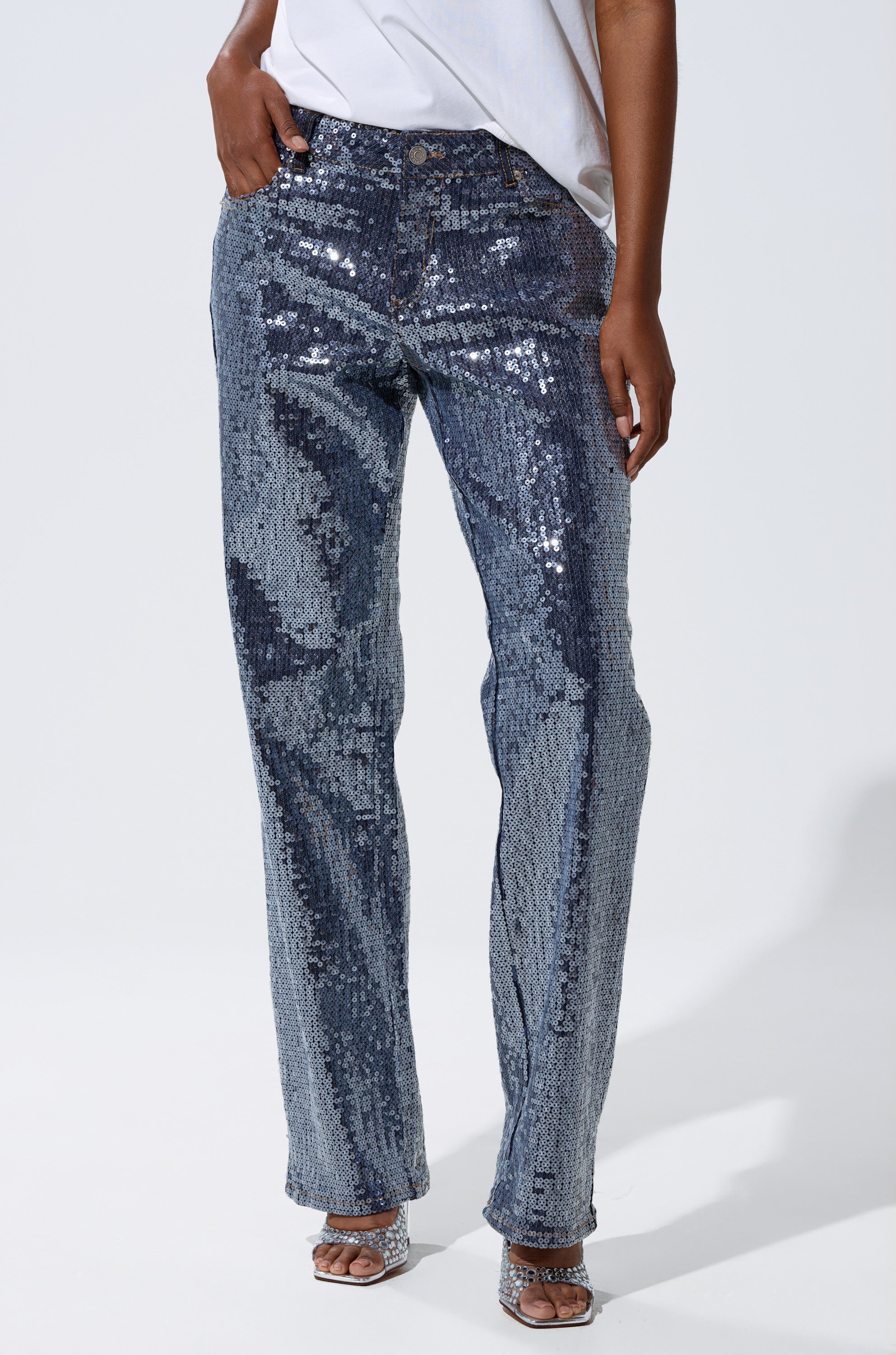 Wearing a white top and ASHTON SEQUIN DENIM PANT with a relaxed fit, one hand in pocket, the person stands in heeled sandals against a plain light background.