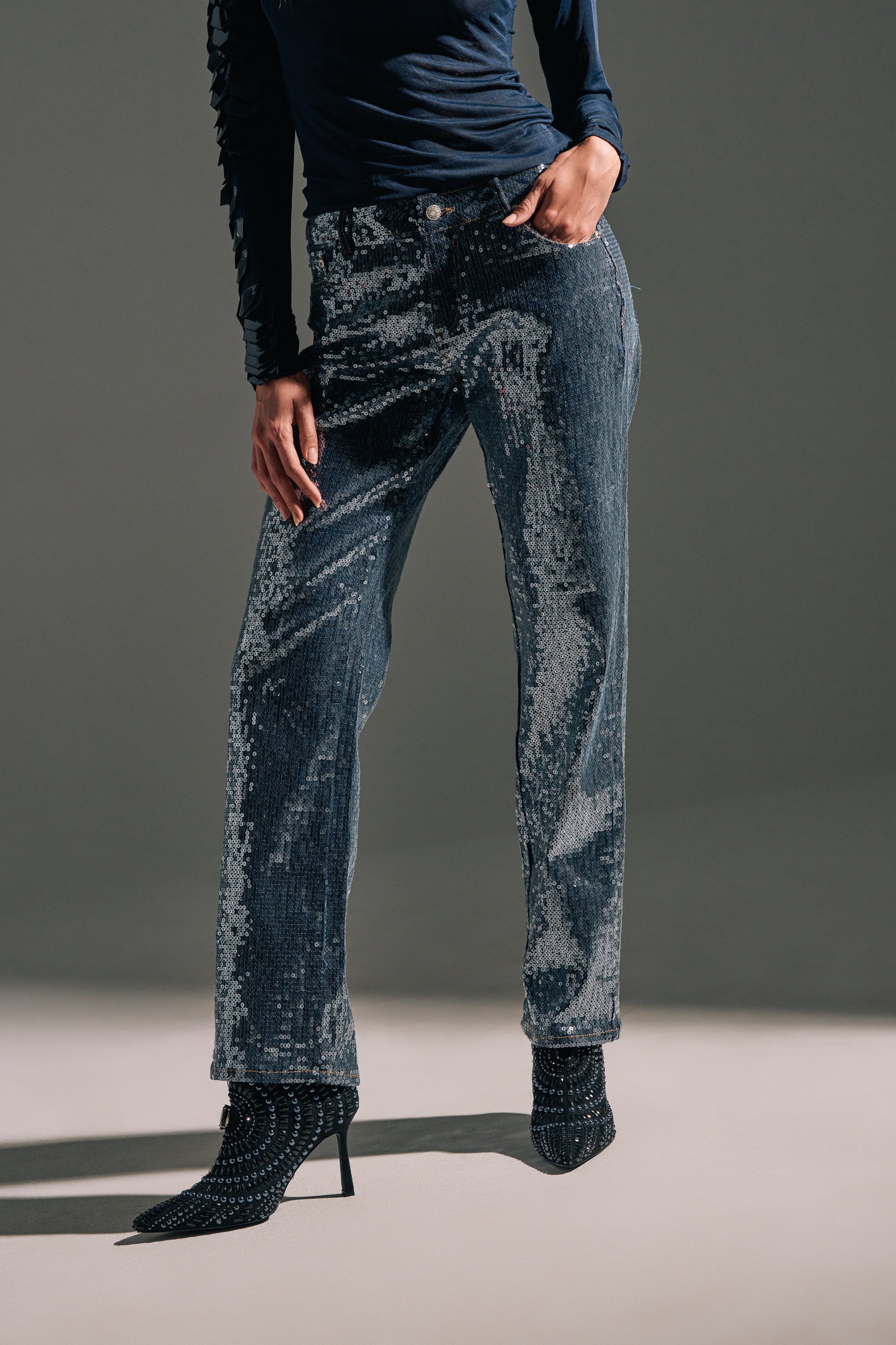 Cropped above the shoulders, a person models the ASHTON SEQUIN DENIM PANT with relaxed fit, paired with high-heeled ankle boots and a long-sleeve navy top, all set against a neutral background to highlight the stylish outfit.