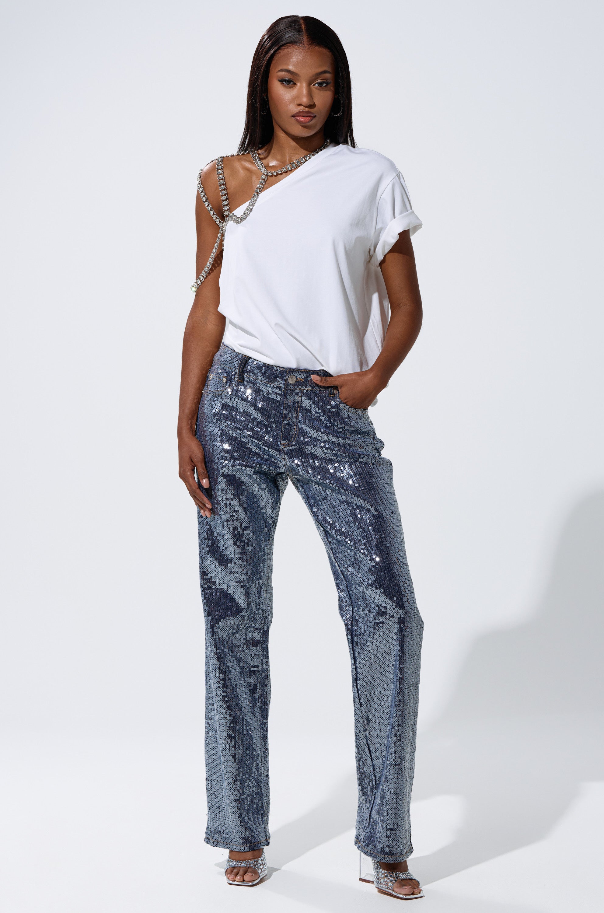 A woman stands confidently in ASHTON SEQUIN DENIM PANT, a white asymmetrical chain-strap t-shirt, and silver heels against a plain white background.