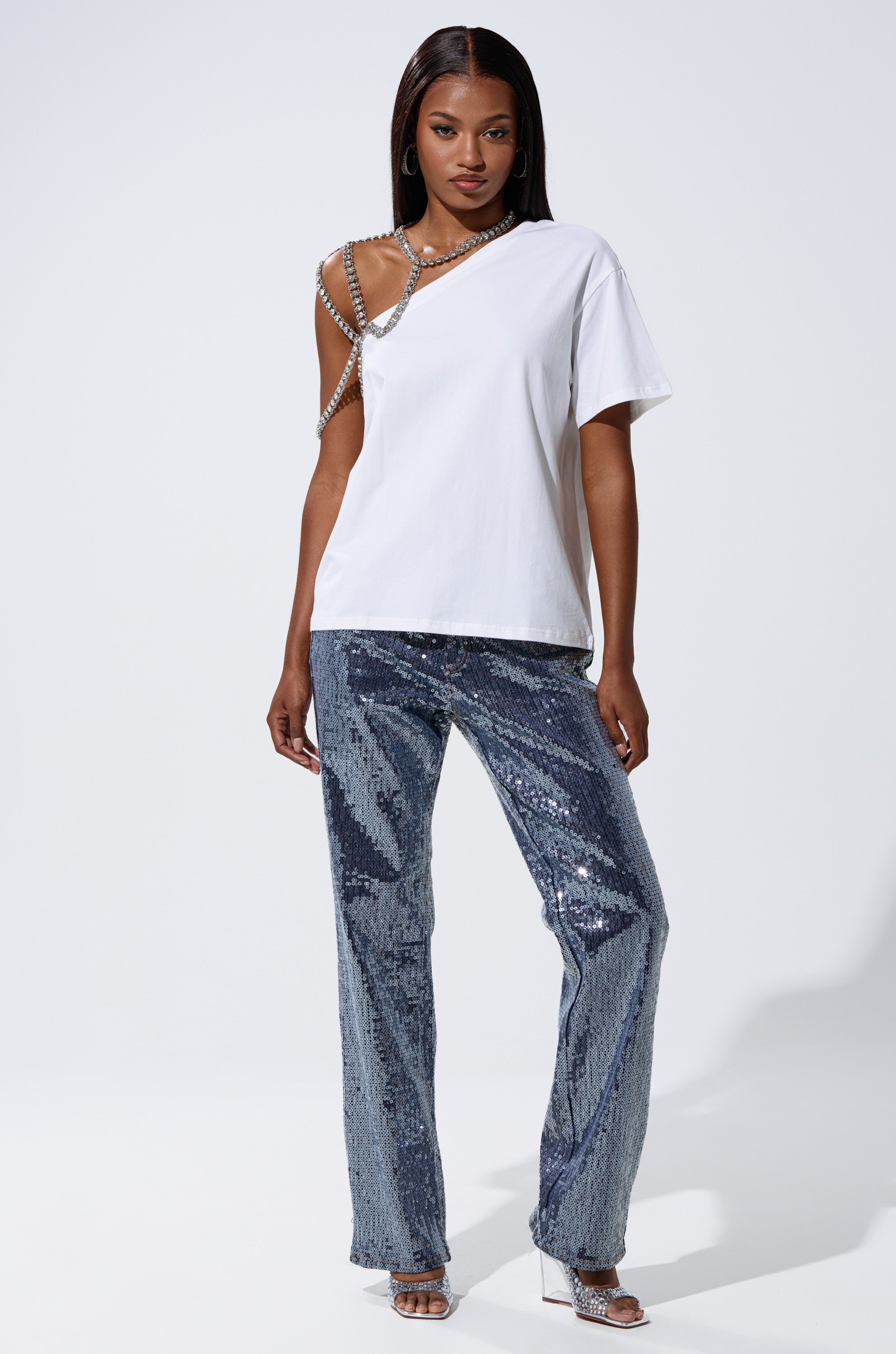 A woman stands against a plain white background wearing the ASHTON SEQUIN DENIM PANT, a white asymmetrical t-shirt with rhinestone strap detail, silver high heels, and minimal jewelry.