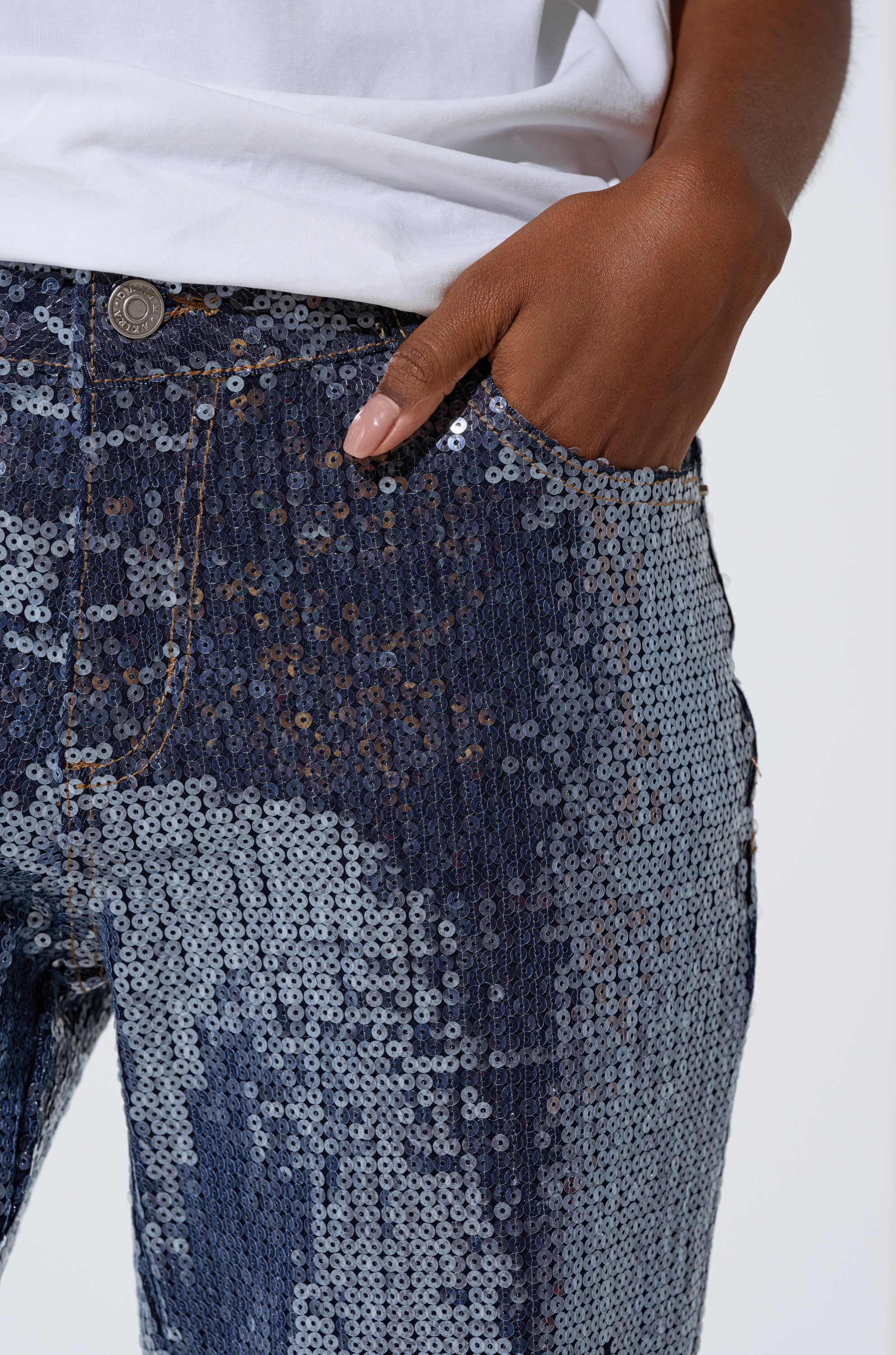 A person wears the ASHTON SEQUIN DENIM PANT and a white shirt, standing with one hand in their pocket to highlight the shiny, reflective texture of these high-rise sequin jeans up close.