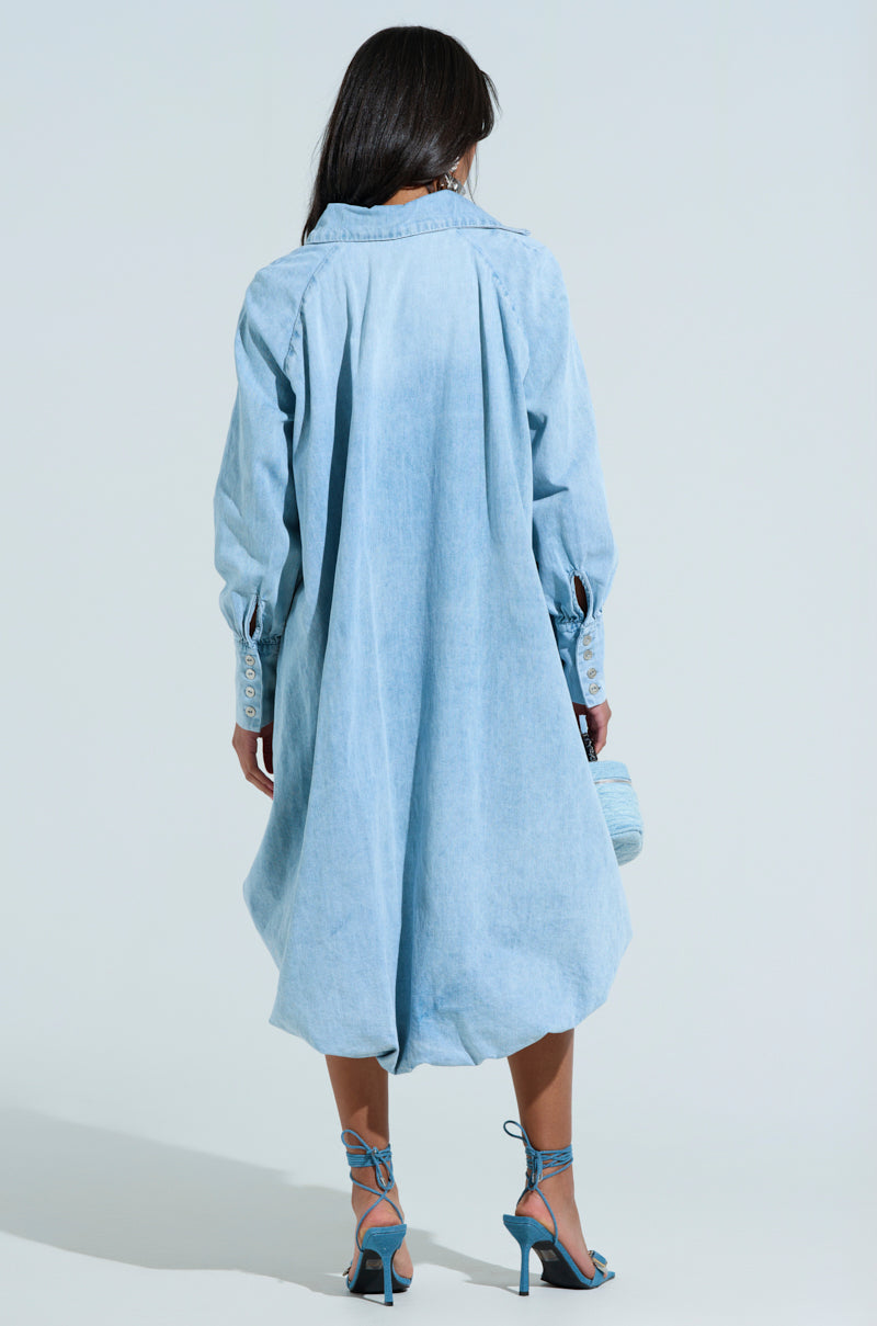 A person with long dark hair is wearing an Amelia Denim High-Low Dress, paired with blue high-heeled sandals and a matching purse. They stand against a plain light grey background, showcasing the dress's chic oversized fit as they face away.