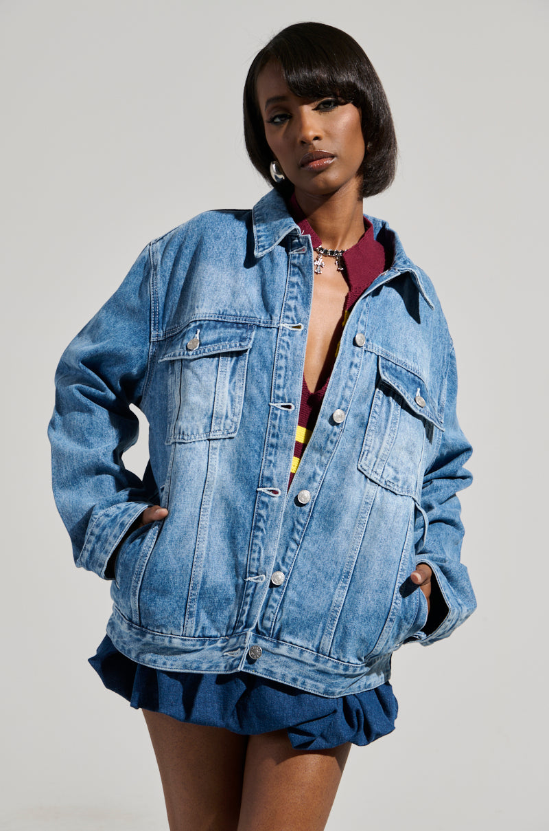 With assured poise, a person dons the GOTTA GO OVERSIZED DENIM JACKET adorned with faux silver hardware over a burgundy top, complemented by a blue ruffled skirt. This medium wash denim ensemble exudes a stylish yet casual vibe against a minimalist gray background.