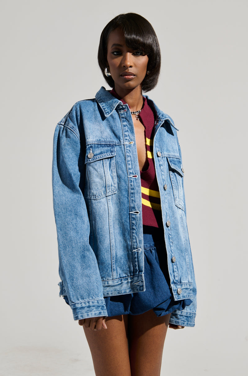 A person stands against a plain gray background, wearing the GOTTA GO OVERSIZED DENIM JACKET with faux silver hardware over a maroon and yellow-striped top and blue shorts. They have a short, sleek hairstyle and maintain a neutral expression. The medium wash denim adds a touch of casual elegance to the ensemble.
