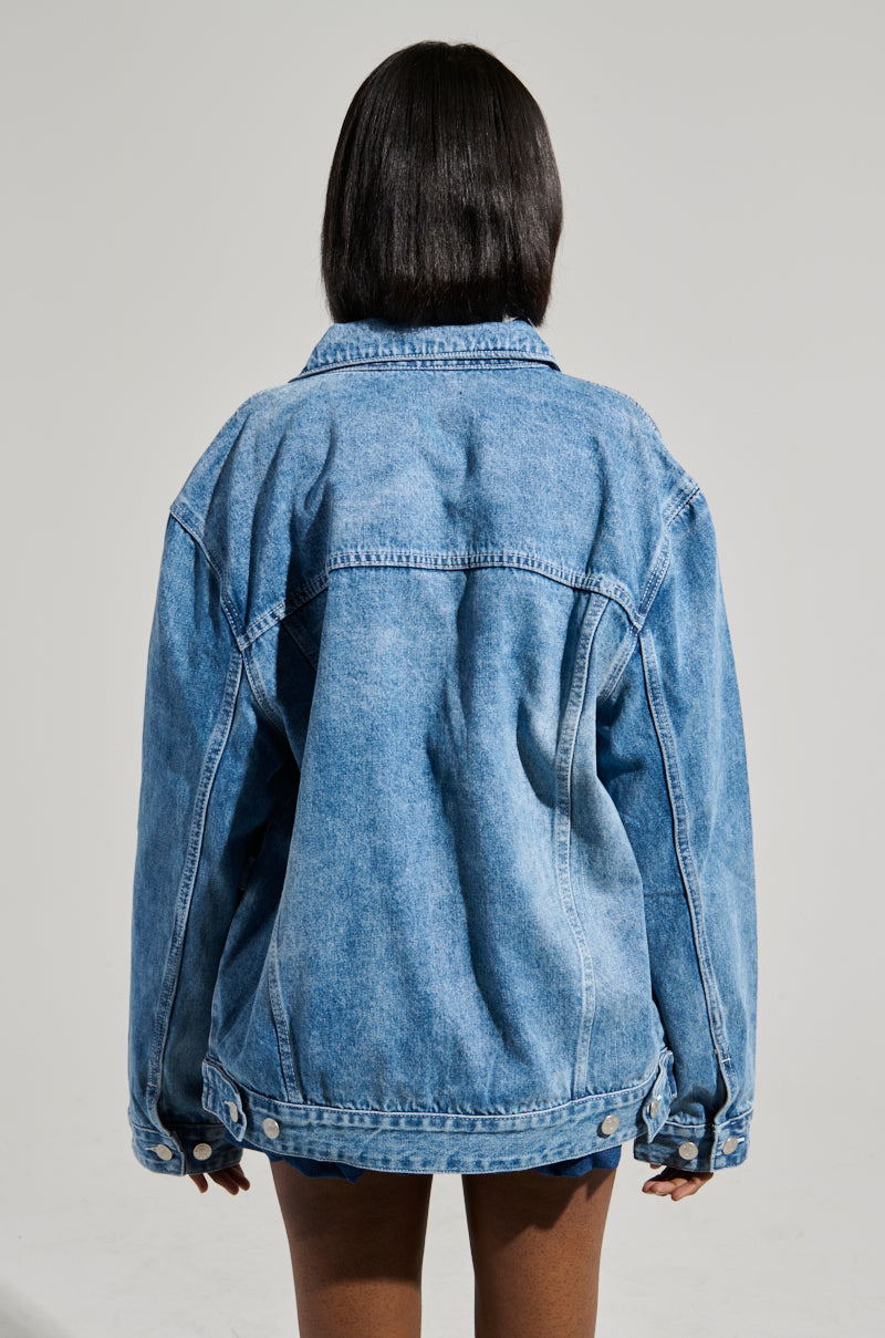 A person with medium-length dark hair is shown from the back wearing the GOTTA GO OVERSIZED DENIM JACKET. The medium wash denim and faux silver hardware pop against the plain, neutral background, emphasizing the jacket's texture and color.
