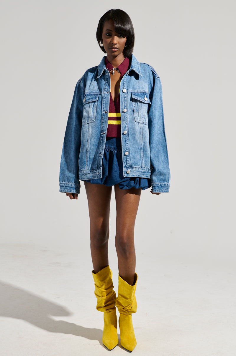 A person stands in a studio wearing the GOTTA GO OVERSIZED DENIM JACKET in medium wash over a burgundy top with yellow and red stripes, paired with blue shorts and distinctive yellow slouchy boots. The background is plain and gray.