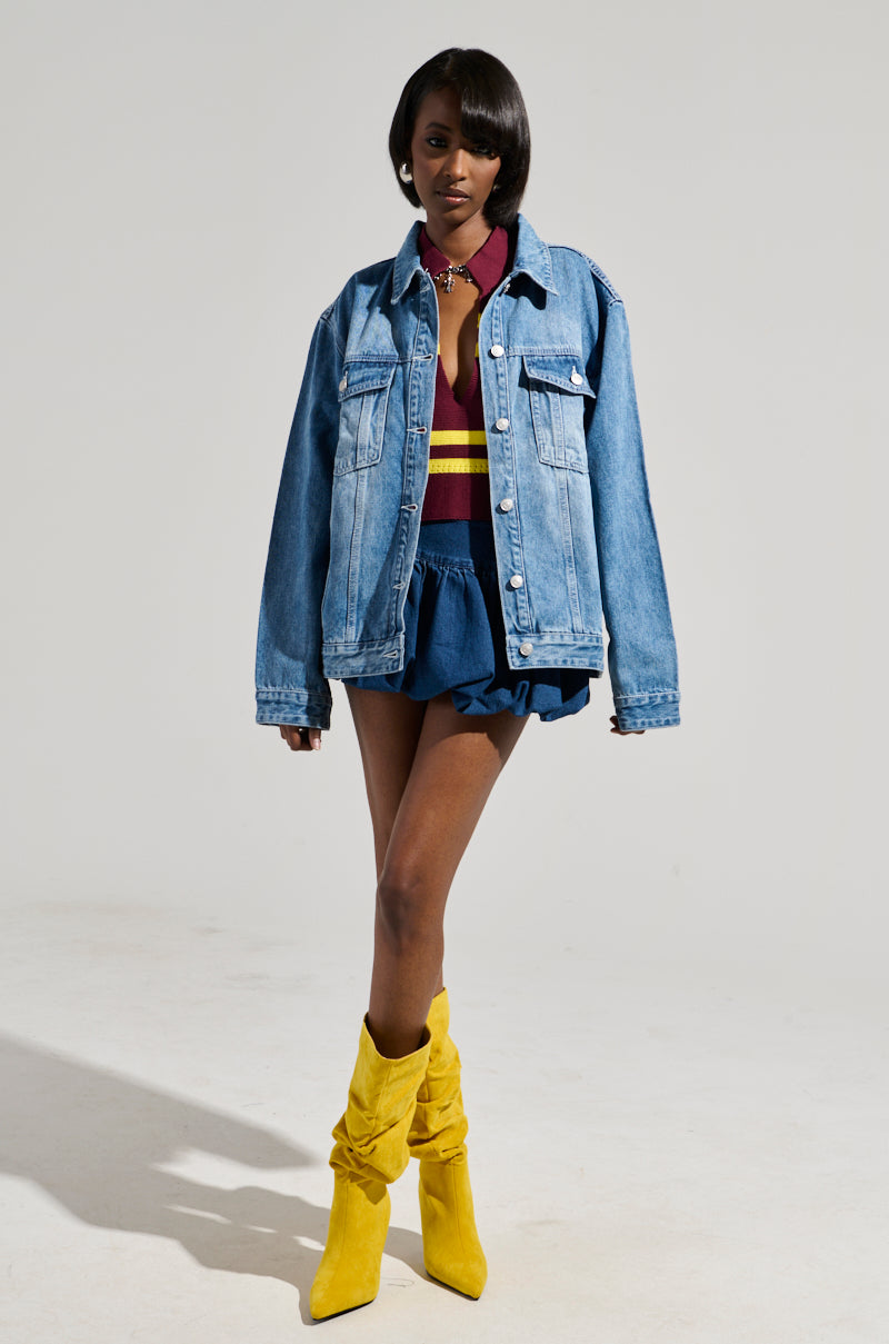 A person poses confidently in the GOTTA GO OVERSIZED DENIM JACKET, featuring medium wash denim and faux silver hardware, layered over a maroon and yellow top with a blue skirt. They complete the fashion-forward look with bright yellow slouchy boots against a plain background, enhancing their stylish ensemble.