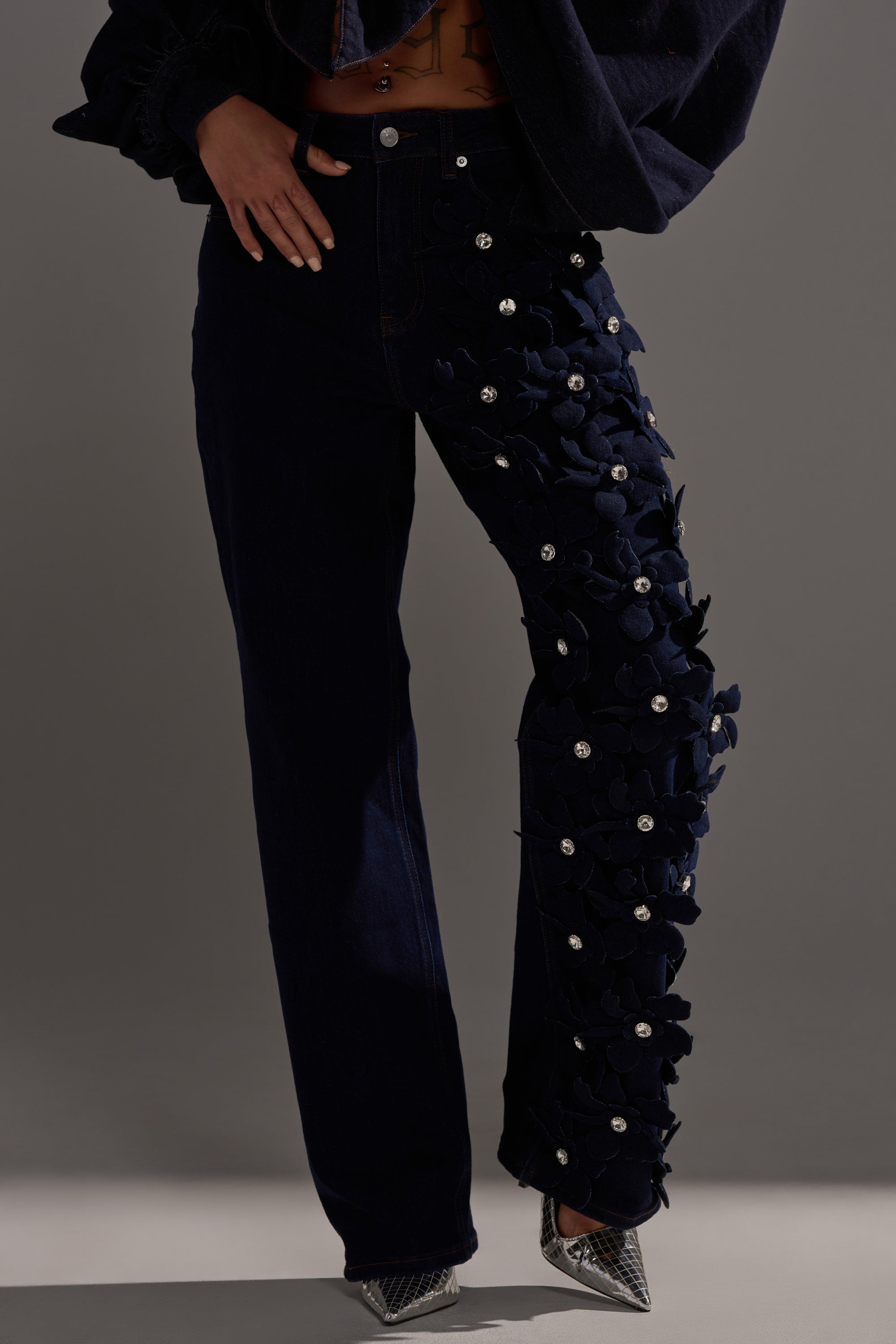 Someone wearing FLORAL FIELDS APPLIQUE DENIM PANTS WITH RHINESTONES, which feature 3D floral details and rhinestone embellishments on one leg, pairs them with silver pointed-toe shoes and a dark jacket against a plain background.