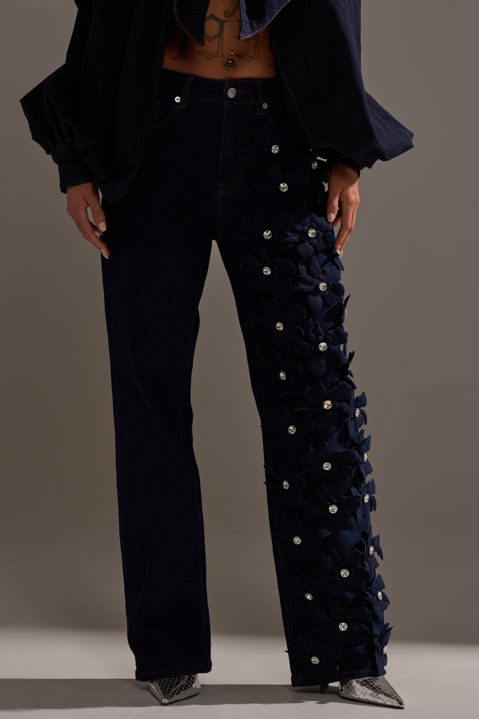 A person wears the FLORAL FIELDS APPLIQUE DENIM PANTS WITH RHINESTONES, featuring a plain right leg and a left leg adorned with dark floral appliqué and rhinestones, paired with a navy top and pointed silver shoes.