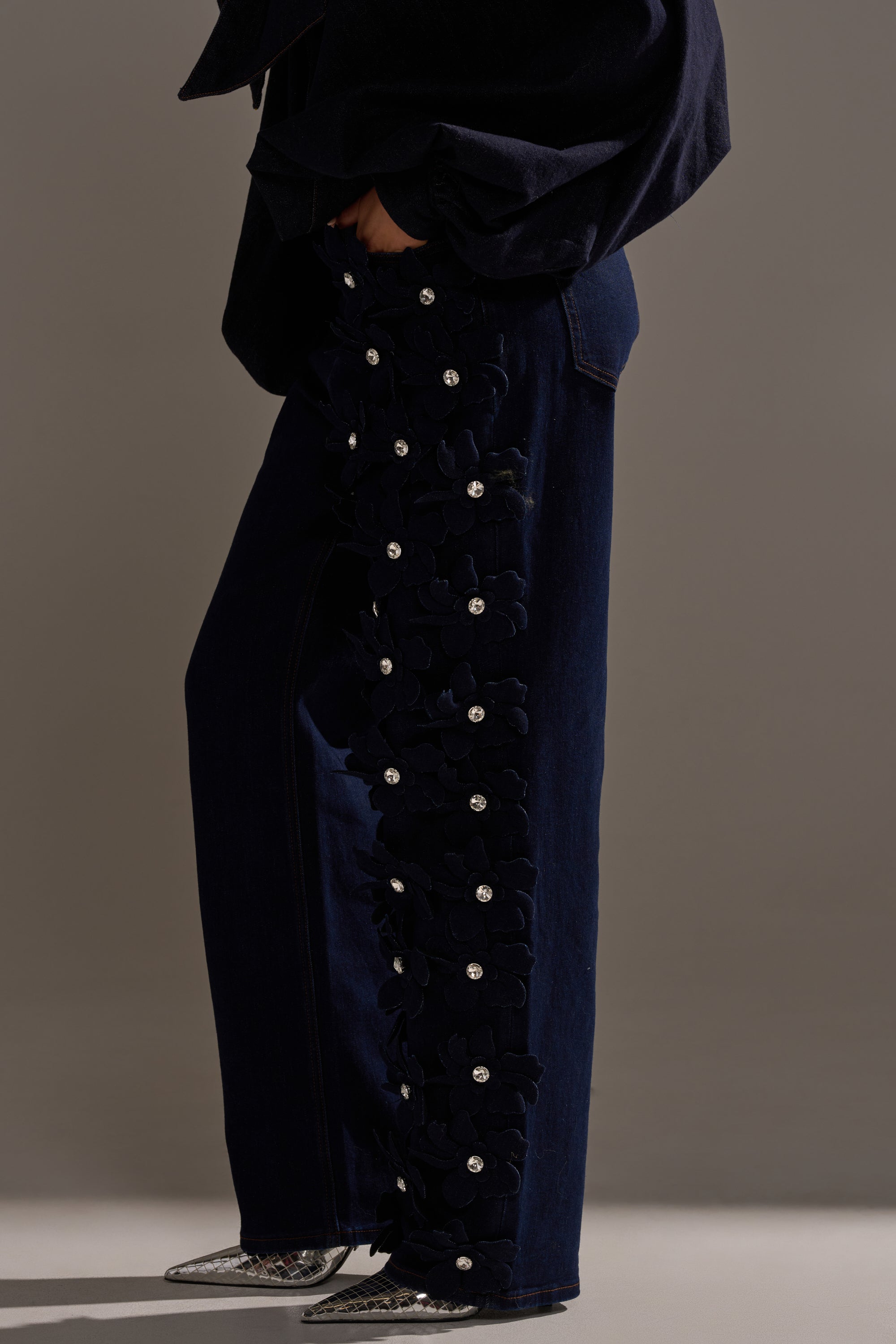 Wearing FLORAL FIELDS APPLIQUE DENIM PANTS WITH RHINESTONES, featuring bold black floral appliqués and rhinestone-embellished side seams, styled with pointed silver shoes and a loose dark top for a striking look.