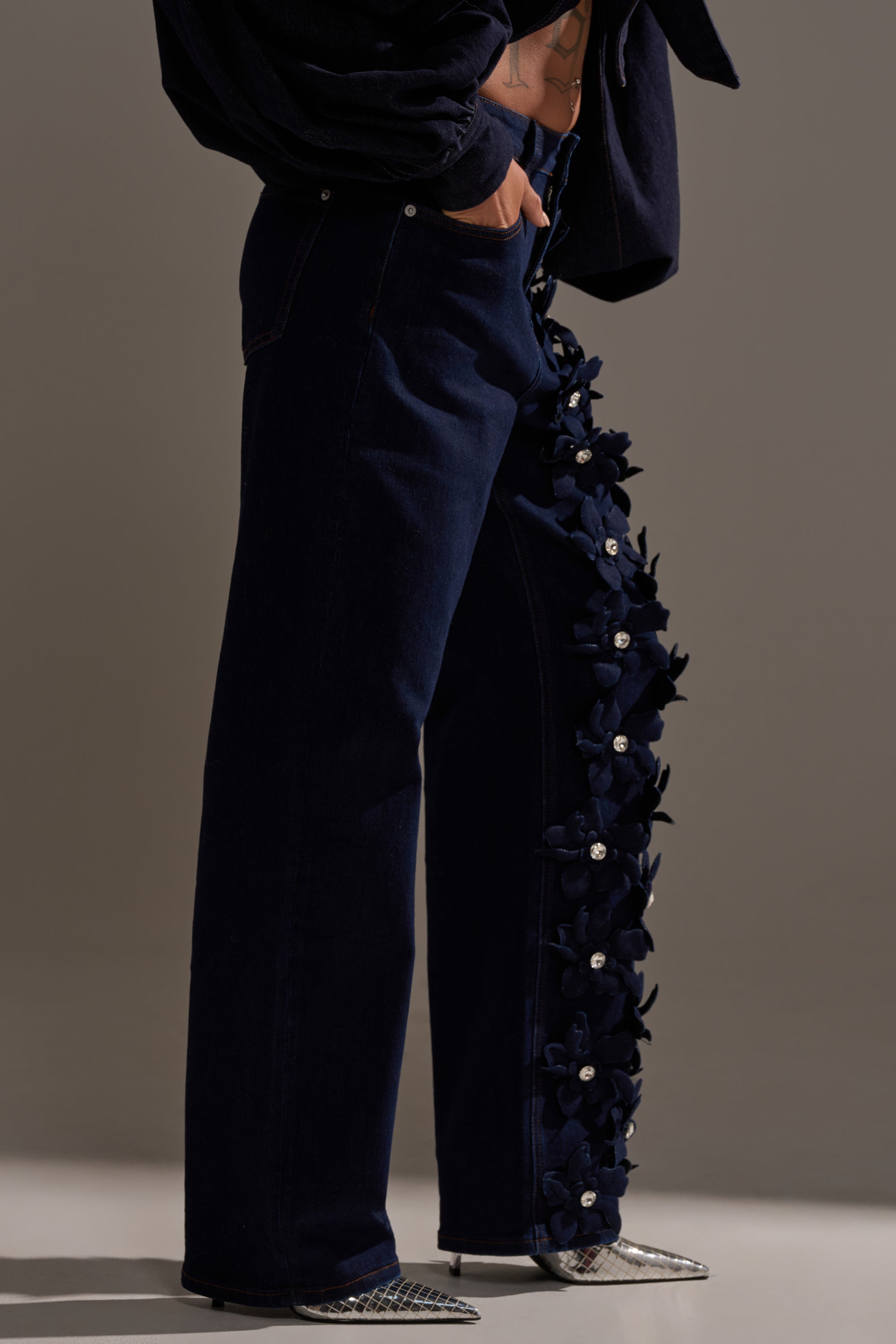 Wearing FLORAL FIELDS APPLIQUE DENIM PANTS WITH RHINESTONES, silver pointed shoes, and a dark top, a person poses with one hand in their pocket against a neutral background.