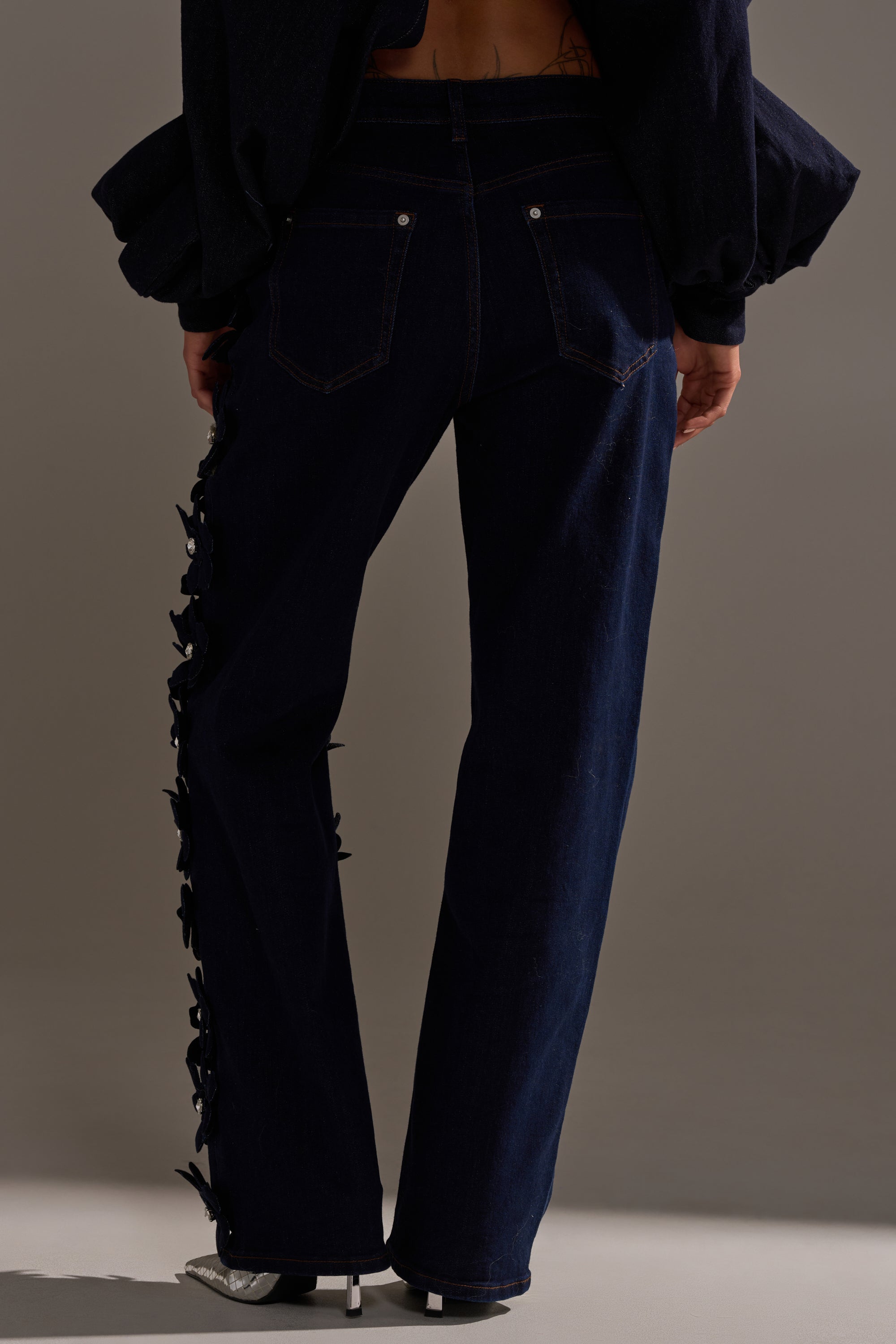 A person faces away wearing FLORAL FIELDS APPLIQUE DENIM PANTS WITH RHINESTONES, featuring high-rise straight legs with black floral embellishments along the seams, paired with a dark top and silver open-toe heels.