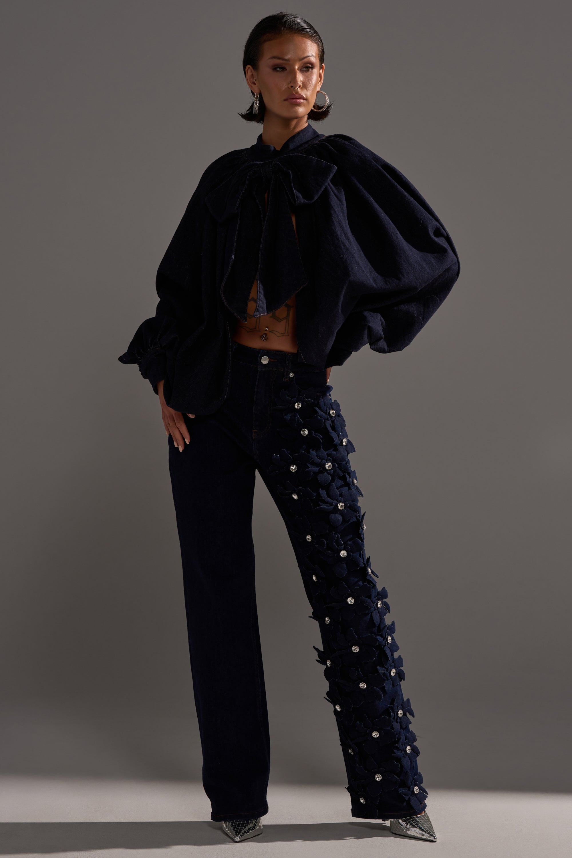 A person poses in a dramatic dark outfit with a voluminous cape and FLORAL FIELDS APPLIQUE DENIM PANTS WITH RHINESTONES, standing against a plain gray background.
