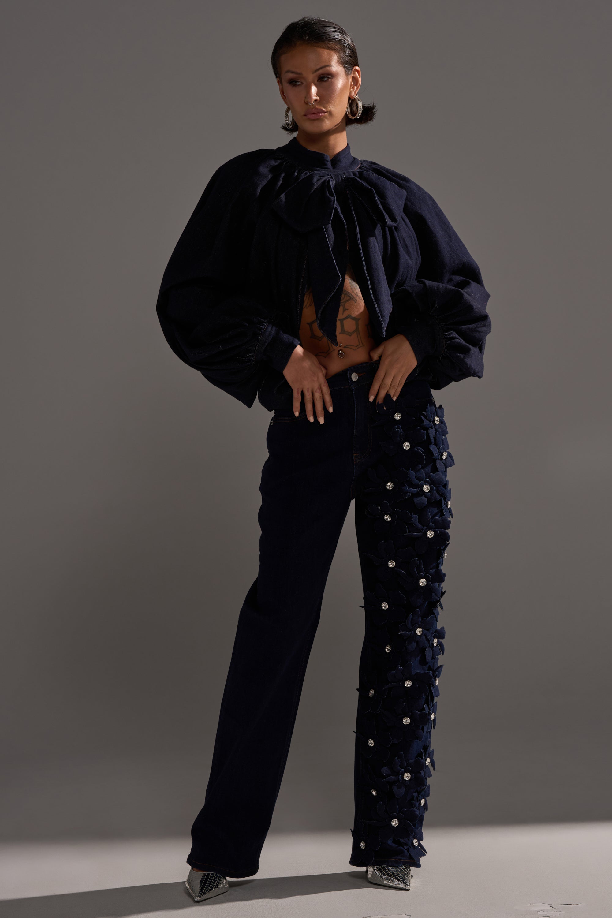 A woman with short hair and statement earrings poses against a gray background, wearing a navy blouse with voluminous sleeves and a bow at the neck, paired with FLORAL FIELDS APPLIQUE DENIM PANTS WITH RHINESTONES featuring textured embellishments.