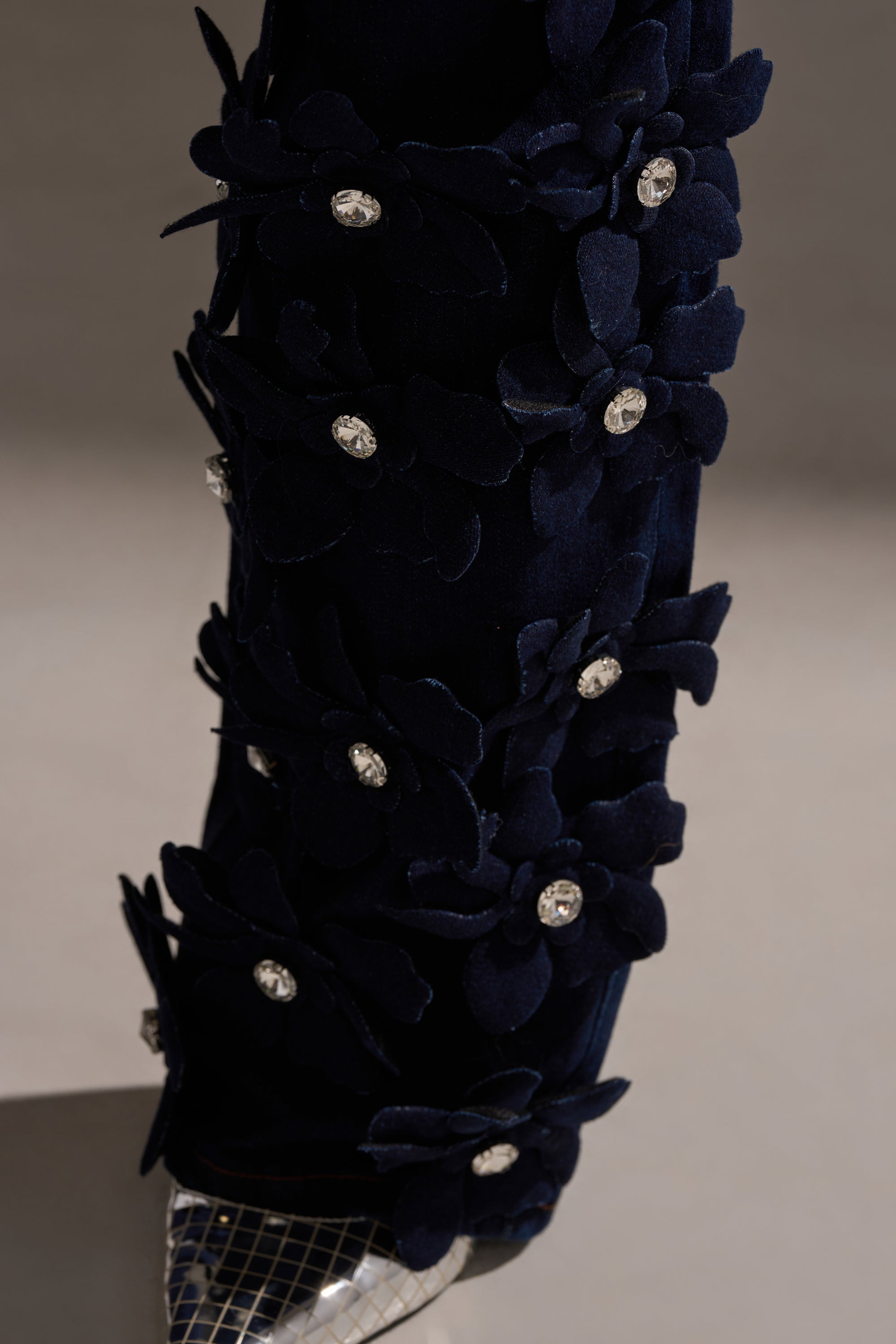 Close-up of the FLORAL FIELDS APPLIQUE DENIM PANTS WITH RHINESTONES, featuring textured floral appliqués and sparkling rhinestones on dark straight-leg denim, with a hint of a metallic mesh shoe visible at the hem.