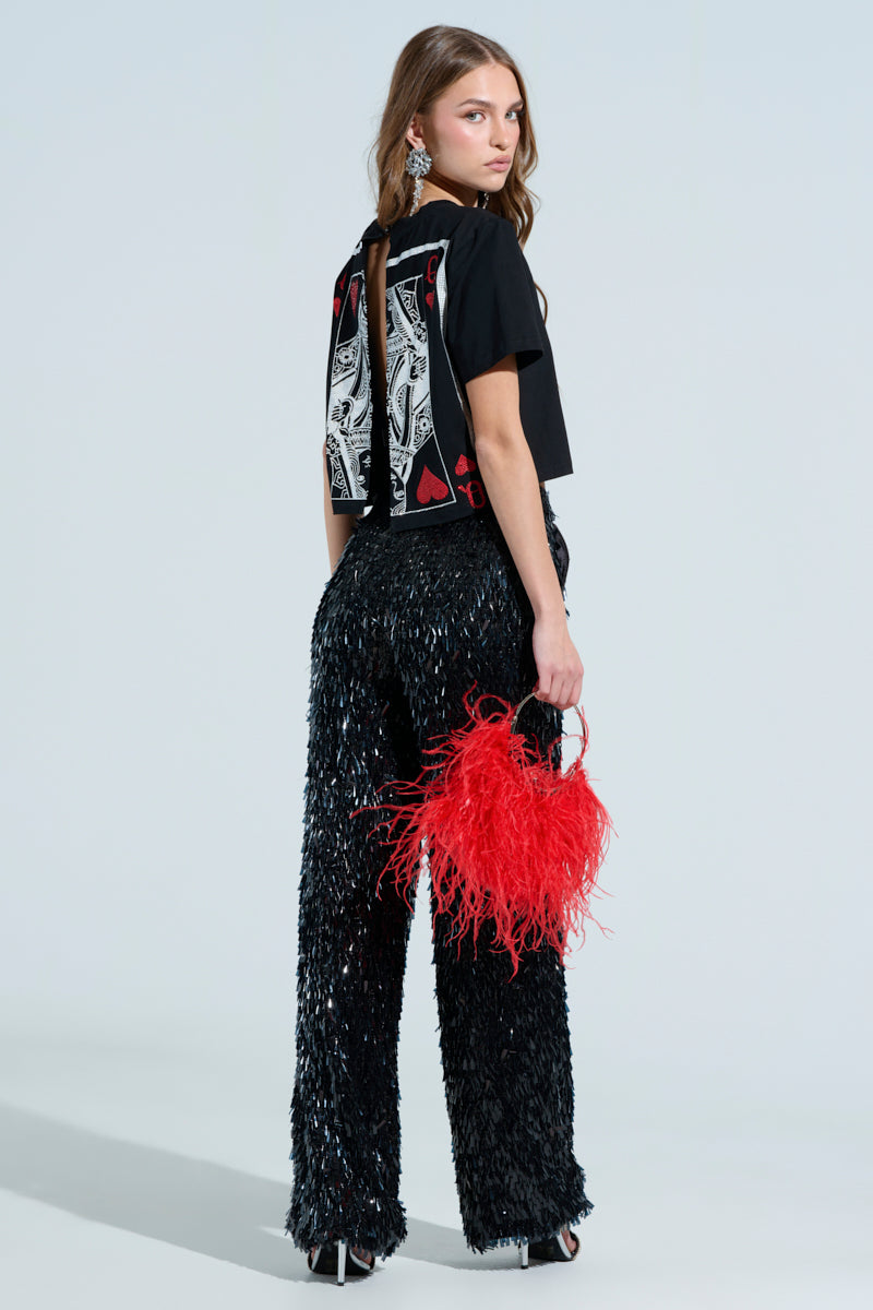 A woman stands sideways wearing a black LUCKY WINNER T-SHIRT featuring a rhinestone-embellished queen of hearts design, paired with shiny black pants. She holds a red feathered handbag, her long hair is loose, and she sports large statement earrings against a plain light background.