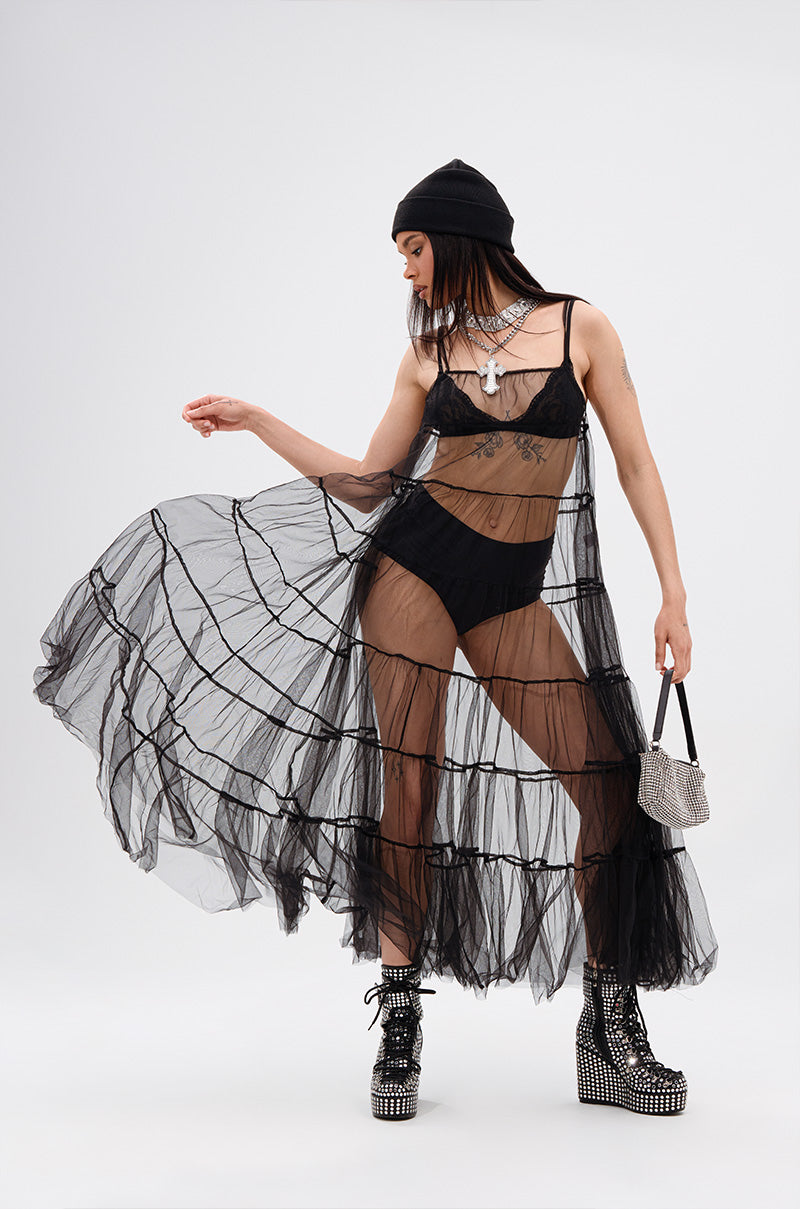 Wearing the PERFECT FOR LAYERING TULLE MAXI DRESS over black lingerie, paired with chunky platform boots, a beanie, layered necklaces, and a small sparkling handbag, a model poses confidently against a plain background.