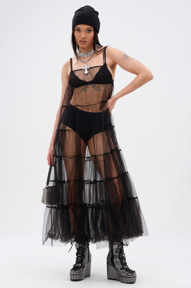 A person stands confidently against a white background wearing the PERFECT FOR LAYERING TULLE MAXI DRESS over black lingerie, styled with a beanie, chunky necklace, studded platform boots, and a small handbag.
