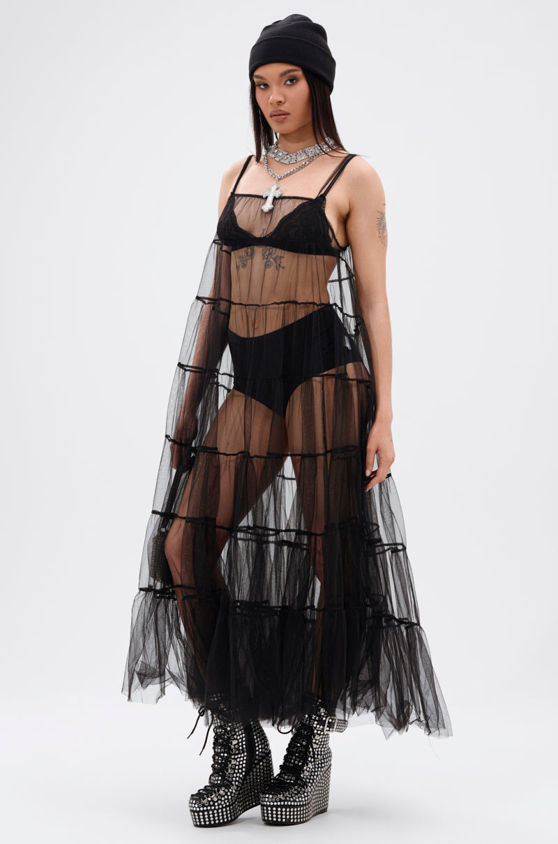 A person stands confidently against a white background wearing the PERFECT FOR LAYERING TULLE MAXI DRESS over black lingerie, paired with studded platform boots, layered necklaces, and a black beanie.