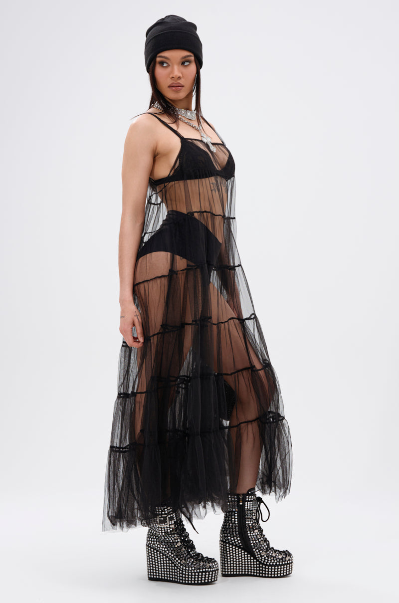 A woman stands sideways against a plain light background in the PERFECT FOR LAYERING TULLE MAXI DRESS, a sheer black maxi with ruffled design over black lingerie, styled with a black beanie, layered necklaces, and studded platform boots.