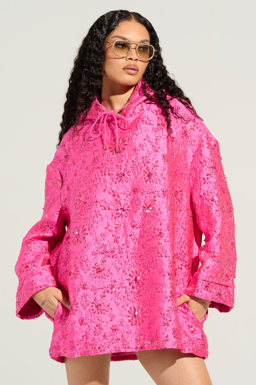 A person with long curly hair and sunglasses is clad in the "WOKE UP LIKE THIS BROCADE SWEATSHIRT DRESS," a bright pink, floral-patterned oversized jacket resembling a hoodie dress, complete with a hood and drawstrings. The jacket is adorned with textured embellishments. The person stands with their hands in the pockets against a neutral background.