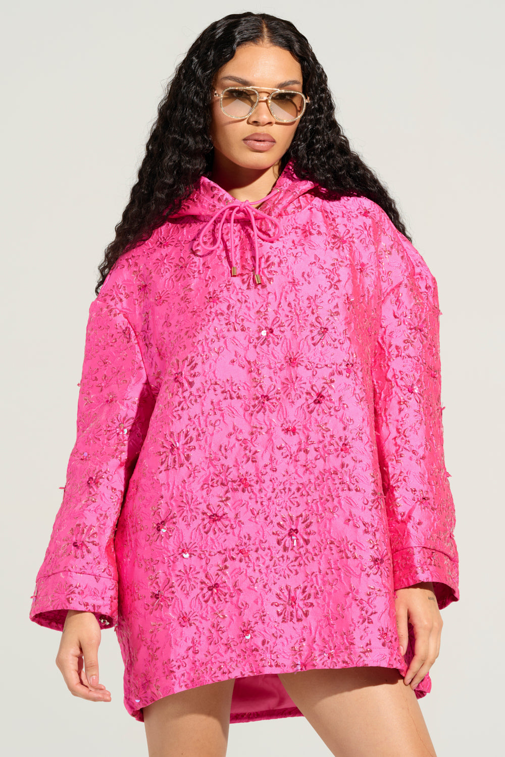 Dressed in the vibrant WOKE UP LIKE THIS BROCADE SWEATSHIRT DRESS—an oversized jacket featuring a rich pink hue with a textured surface and delicate floral design—a person stands confidently. Their long, wavy hair cascades beneath sunglasses, all set against a minimalist light gray backdrop.