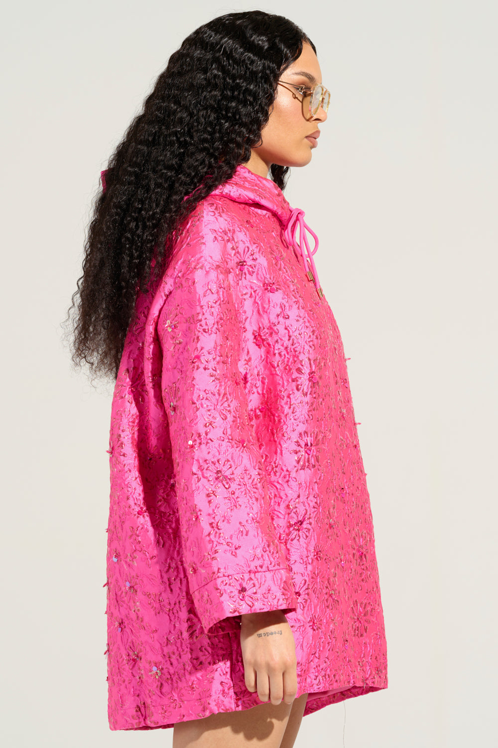 Person wearing the WOKE UP LIKE THIS BROCADE SWEATSHIRT DRESS, a vibrant pink, floral-patterned oversized coat with a hood and drawstrings. They have long curly hair, wear glasses, and have a small tattoo on their inner wrist. The background is a plain, light color reminiscent of brocade fabric.