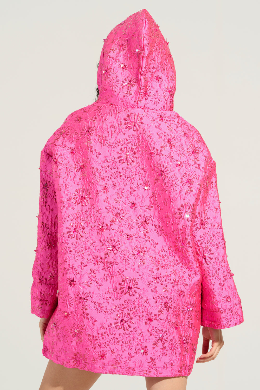 A person wearing the brightly colored WOKE UP LIKE THIS BROCADE SWEATSHIRT DRESS, adorned with floral sequins and brocade-like sparkling details, stands facing away. The oversized hooded garment is accentuated by a plain background, which highlights its vivid and captivating design.