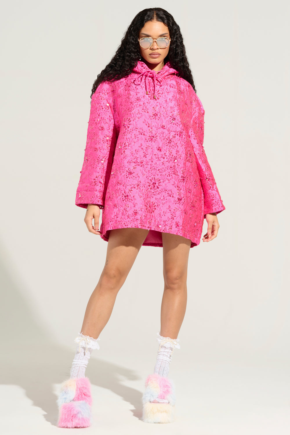 A person with long, curly hair wearing sunglasses, a bright pink WOKE UP LIKE THIS BROCADE SWEATSHIRT DRESS, and white socks with fluffy, multicolored slippers stands against a plain background.