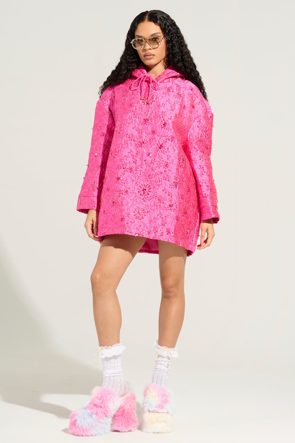 A person in the WOKE UP LIKE THIS BROCADE SWEATSHIRT DRESS, complete with sunglasses and colorful fluffy slippers, stands against a plain background with hands by their sides, exuding a relaxed and stylish vibe.