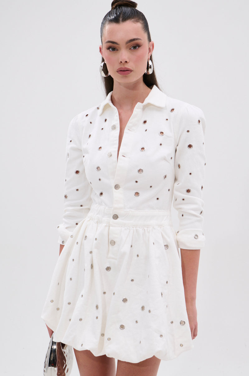 A woman wearing the CUT IT OUT DENIM MINI DRESS WITH GROMMETS stands against a plain background, holding a small metallic handbag. Her long dark hair is styled in a sleek ponytail.