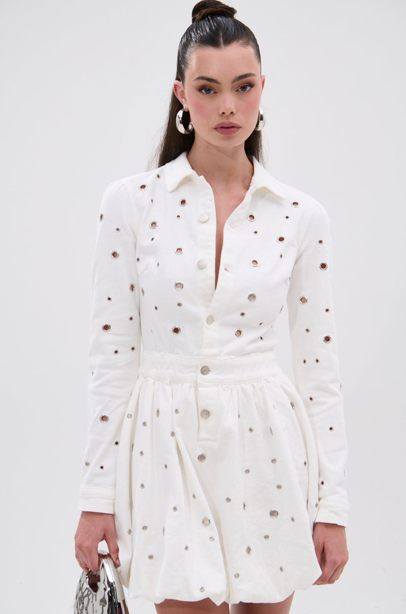 A woman stands against a plain background holding a small bag, wearing the CUT IT OUT DENIM MINI DRESS WITH GROMMETS—featuring metallic grommet details, front buttons, and a gathered waist.