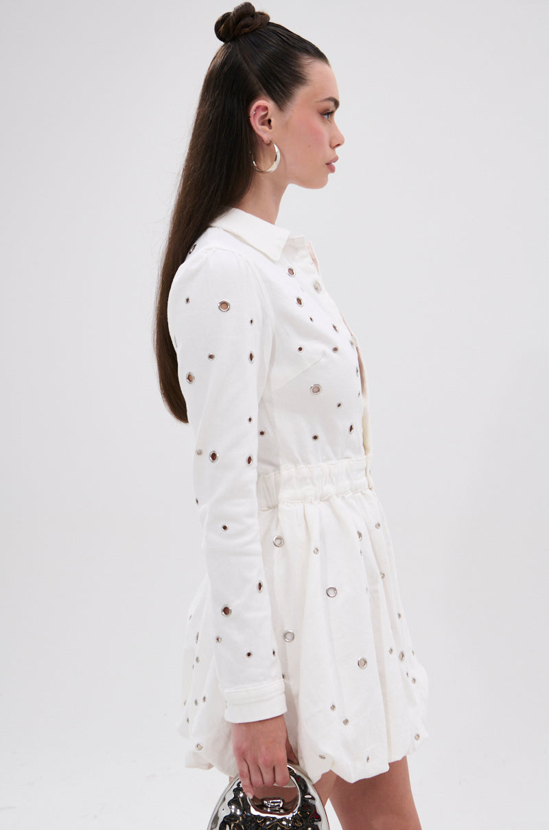 A woman in profile wears the CUT IT OUT DENIM MINI DRESS WITH GROMMETS, featuring a white long-sleeve design, collared neckline, grommet hardware, and cinched waist. She holds a silver handbag and has long, straight half-up hair.