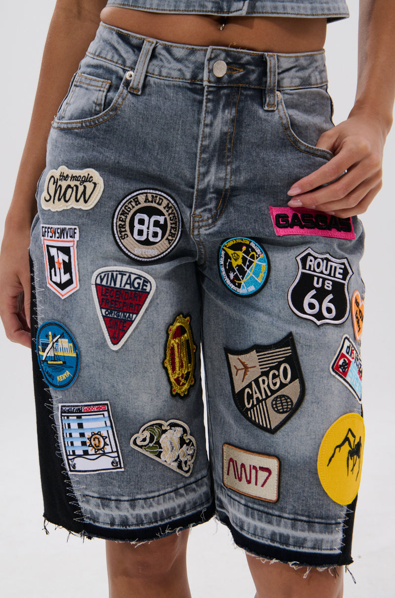 A person wears the VINTAGE DENIM BERMUDA SHORT WITH PATCHES, featuring colorful embroidered badges, logos, and symbols, while standing against a plain background.