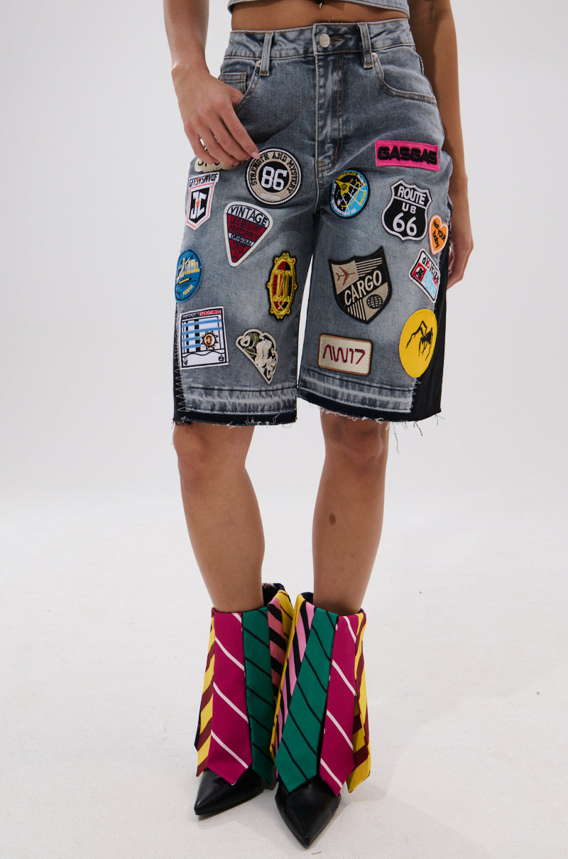 A person wears the VINTAGE DENIM BERMUDA SHORT WITH PATCHES and bold high-heeled boots with pink, yellow, and green diagonal stripes. The photo is cropped at the waist and mid-calf.