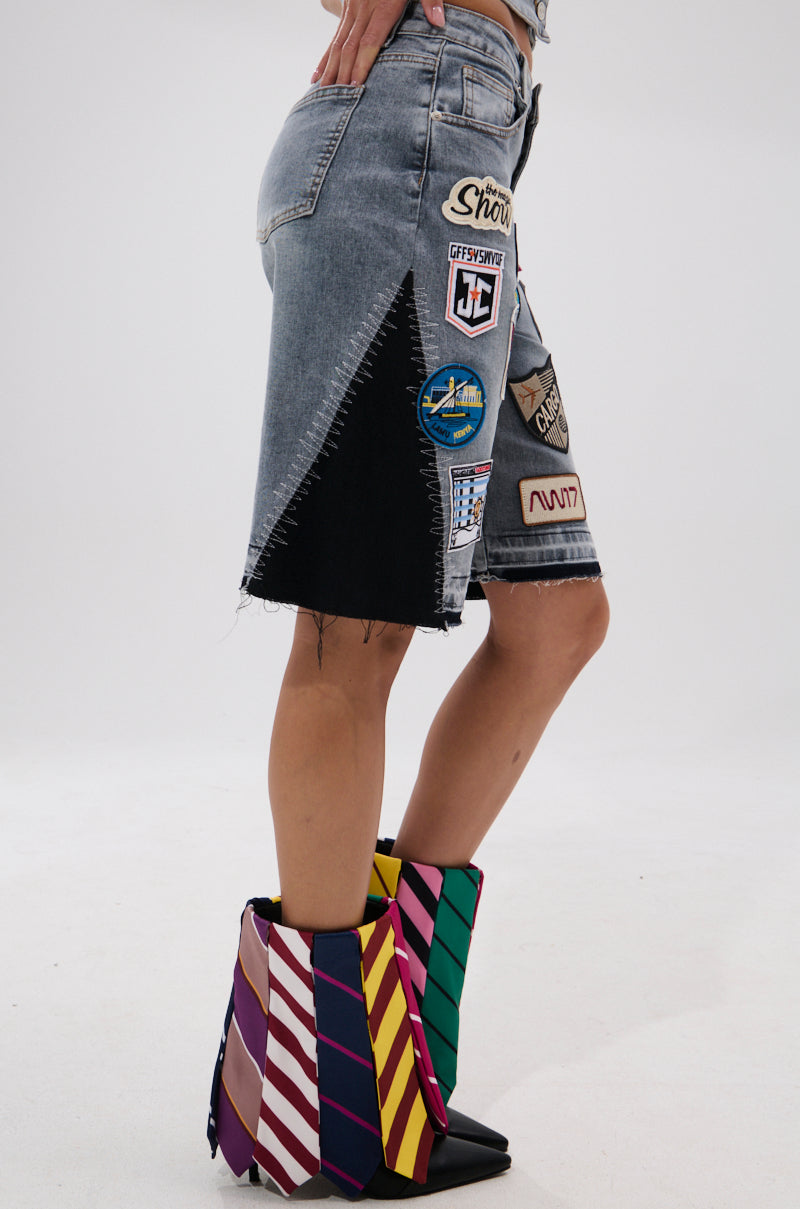 Someone wearing the VINTAGE DENIM BERMUDA SHORT WITH PATCHES, featuring embroidered patches and zigzag-stitched black side fabric, paired with multicolored striped wedge boots.