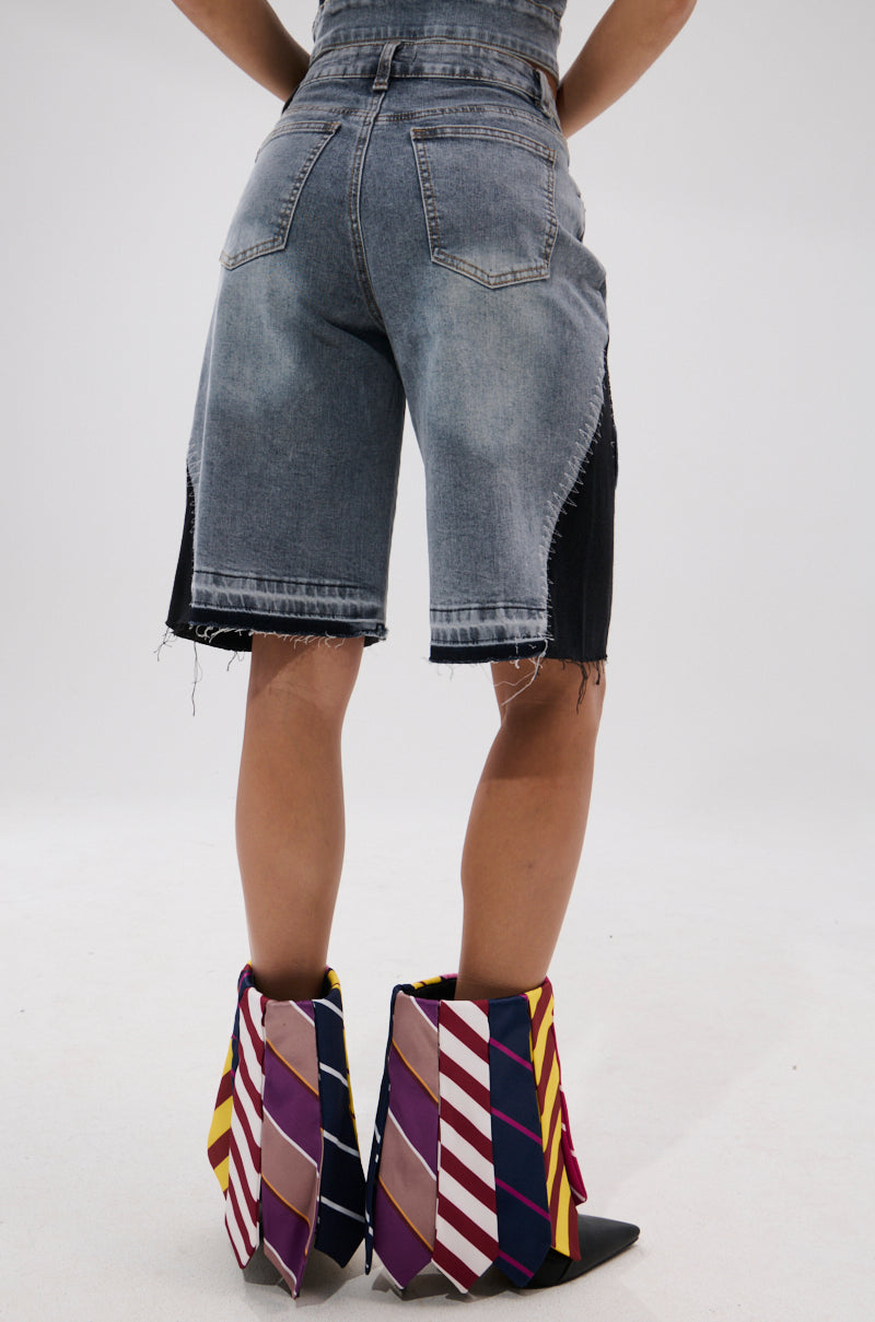 A person stands facing away, wearing VINTAGE DENIM BERMUDA SHORT WITH PATCHES paired with colorful, pointed boots featuring bold diagonal stripes in purple, red, yellow, and white. The background is plain and light-colored.