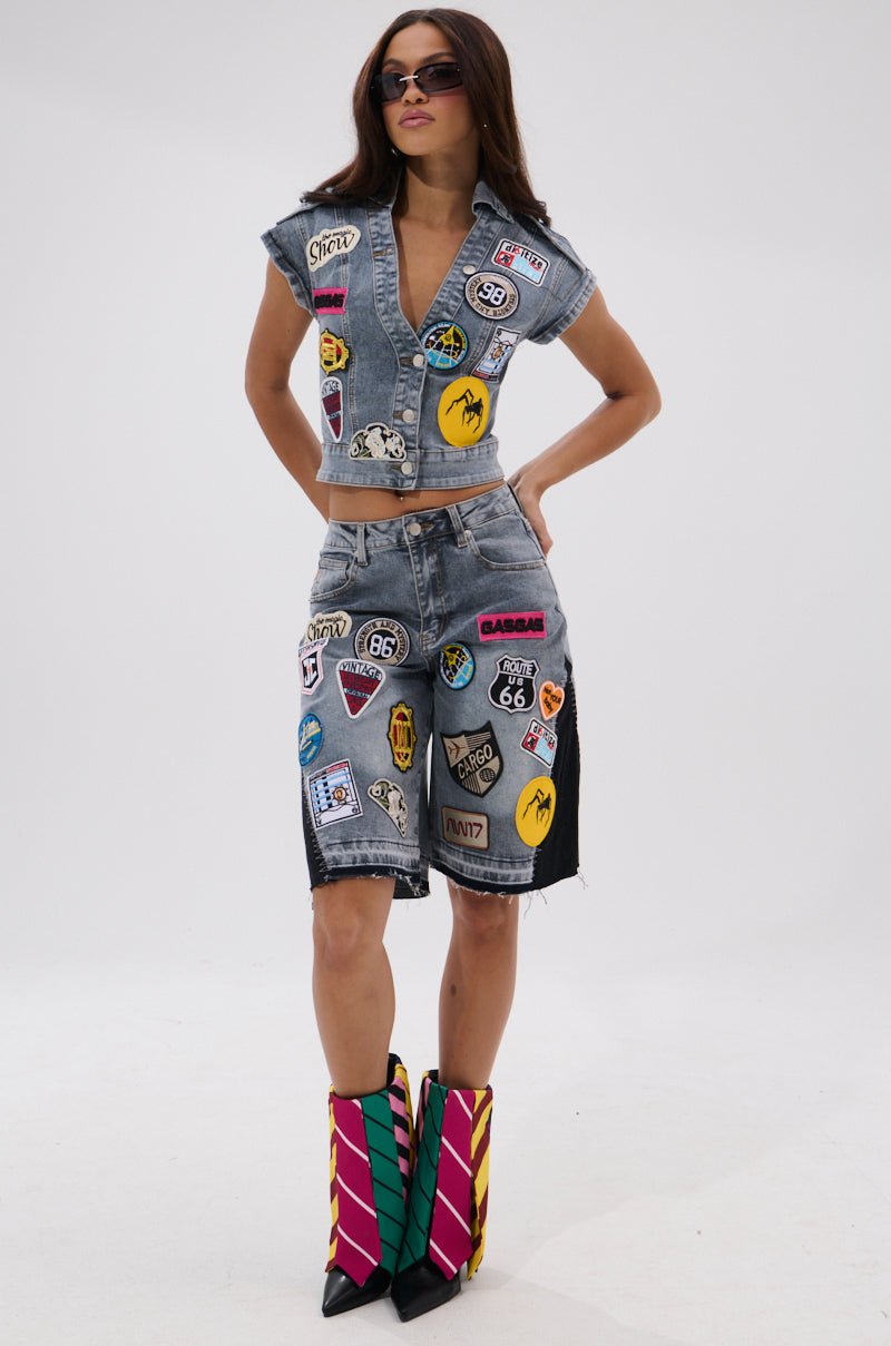 A woman models the VINTAGE DENIM BERMUDA SHORT WITH PATCHES and a matching vest, both detailed with colorful patches, styled with striped pointed boots and sunglasses against a plain white background.