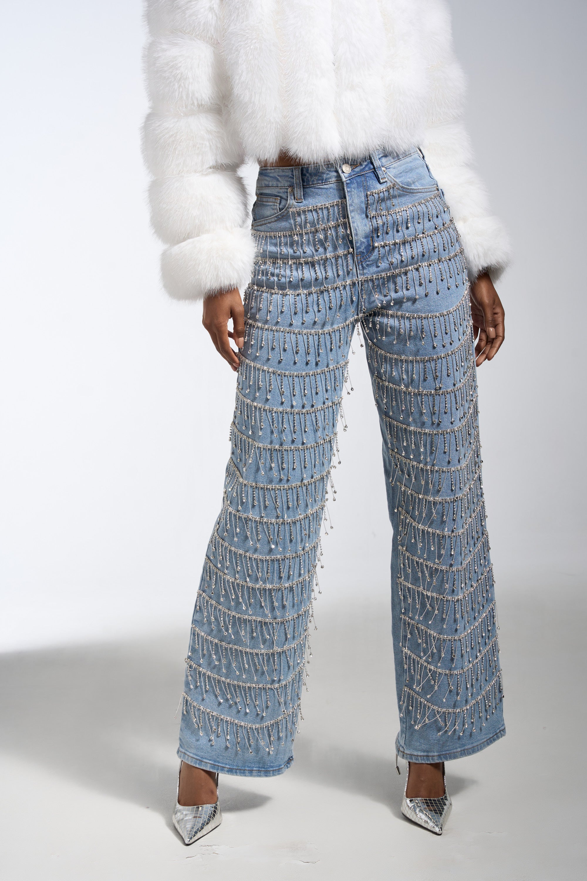 A person stands against a plain background wearing DRIP DRIP EMBELLISHED DENIM high-rise wide-leg jeans with sparkling silver beads, a white furry jacket, and pointed silver high heels.