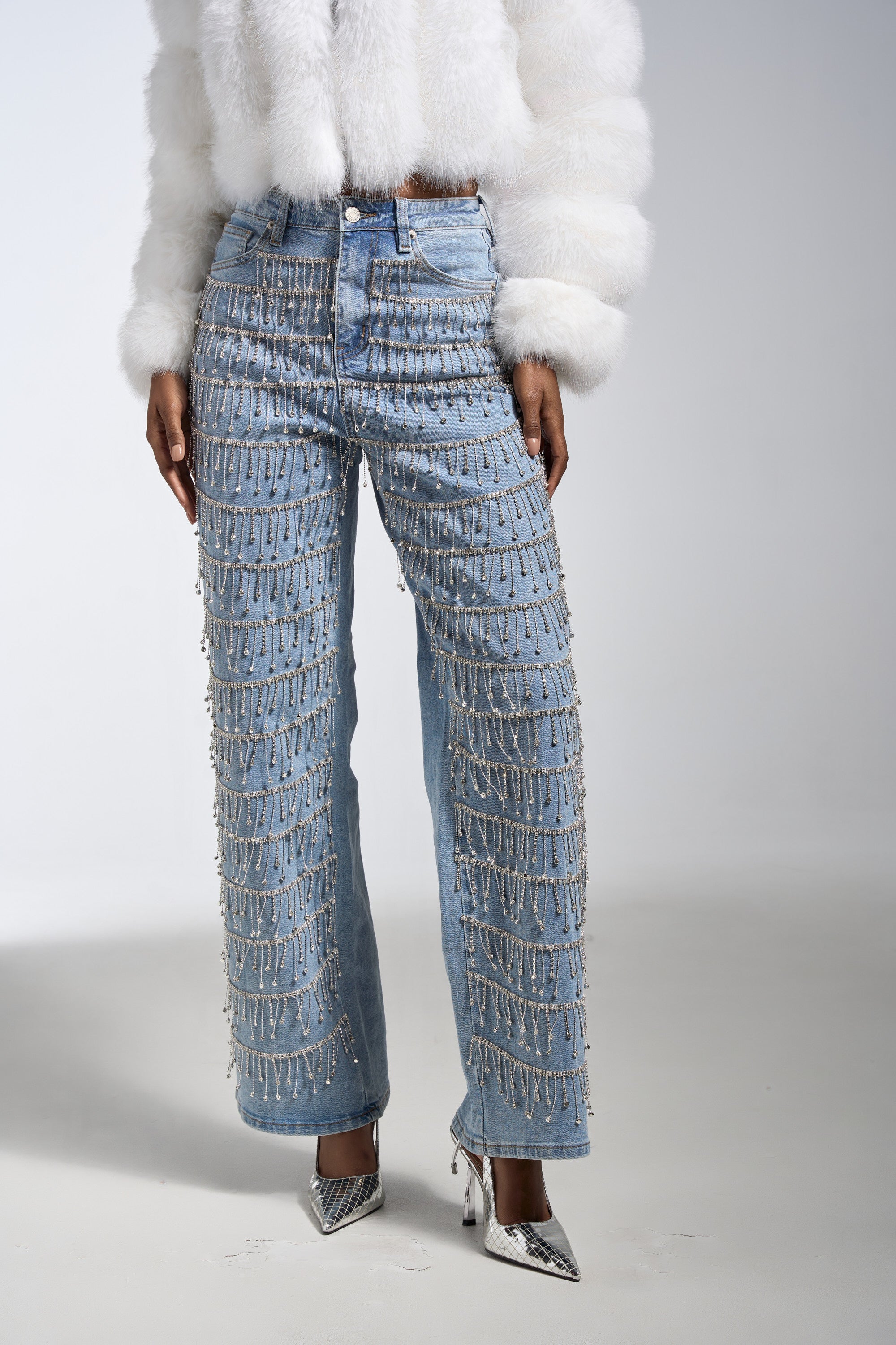 Wearing the DRIP DRIP EMBELLISHED DENIM—high-rise straight-leg jeans adorned with rows of dangling silver beads—a person pairs them with a white faux fur jacket and pointed silver heels, standing against a plain light background.