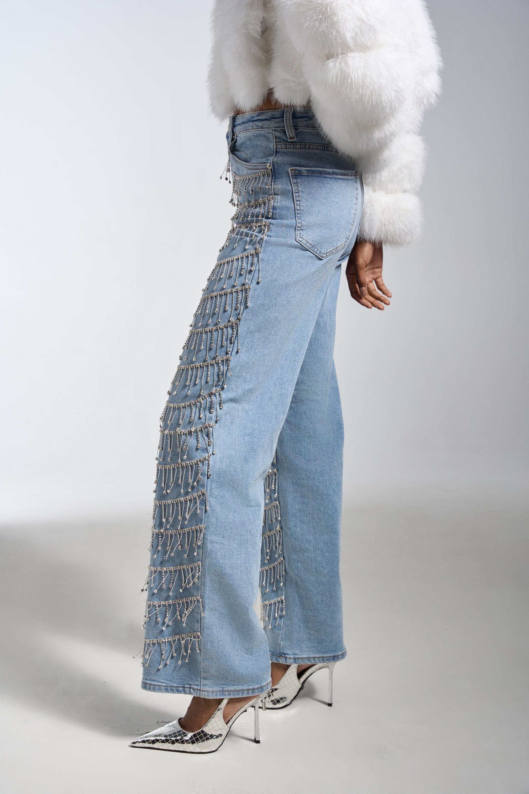 Someone wears the DRIP DRIP EMBELLISHED DENIM in light blue with a white faux fur jacket and pointed silver snakeskin heels, standing against a plain background.