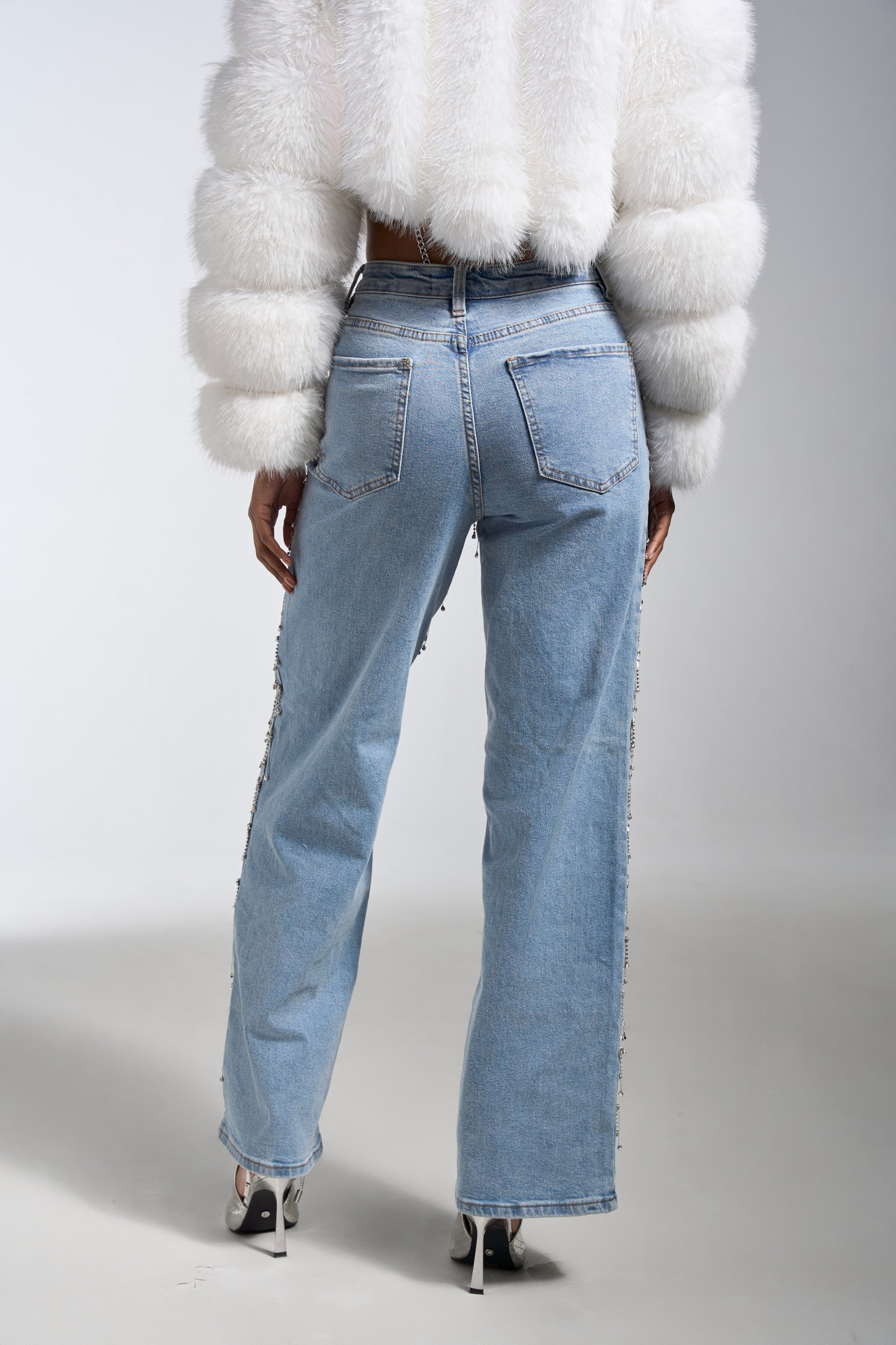 A person photographed from the back wears a cropped white faux fur jacket, DRIP DRIP EMBELLISHED DENIM jeans, and white high-heeled shoes against a plain light background.