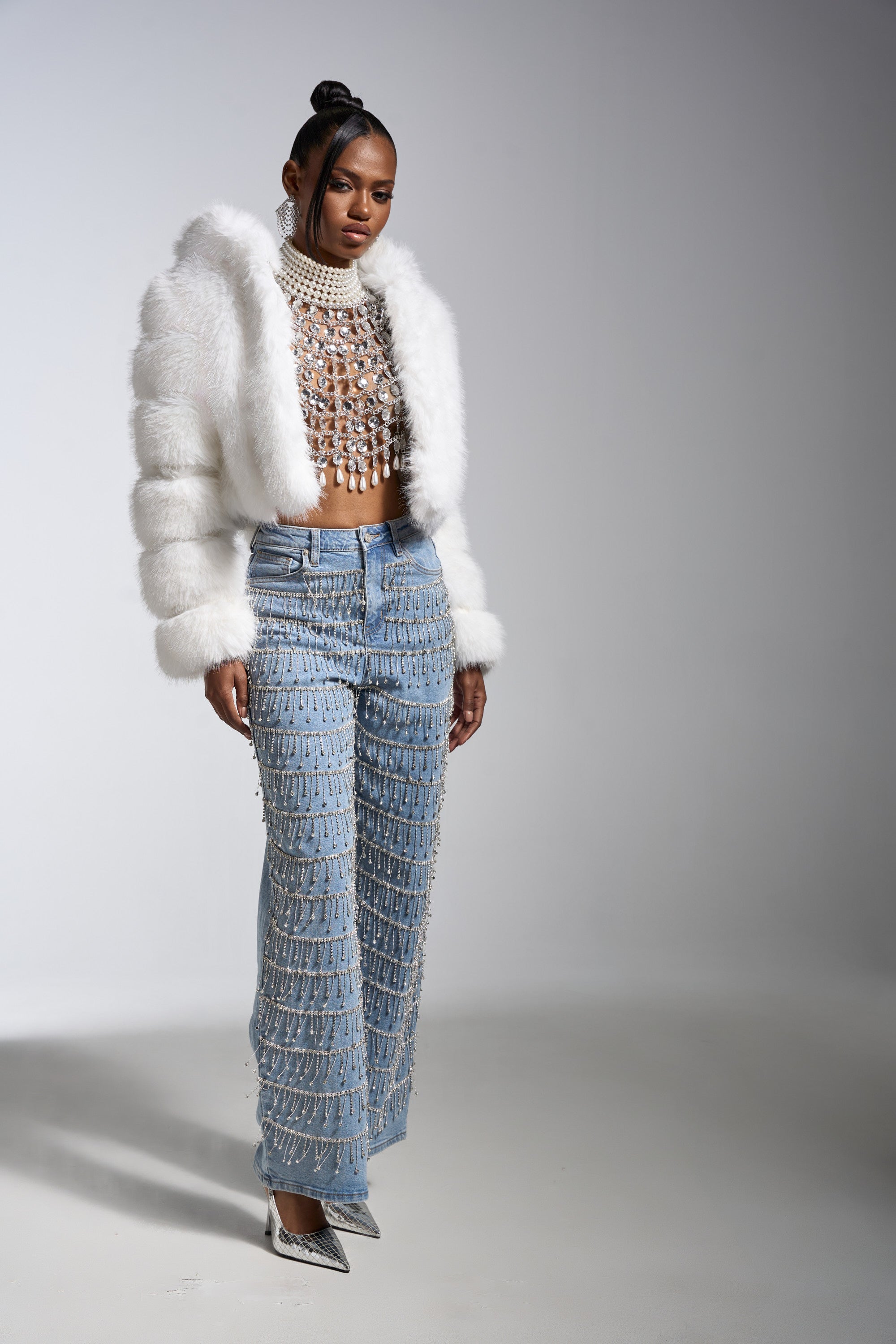 A woman stands in a studio wearing the DRIP DRIP EMBELLISHED DENIM jeans, paired with a white faux fur jacket, beaded crop top, silver pointed heels, and her hair styled in a sleek bun.